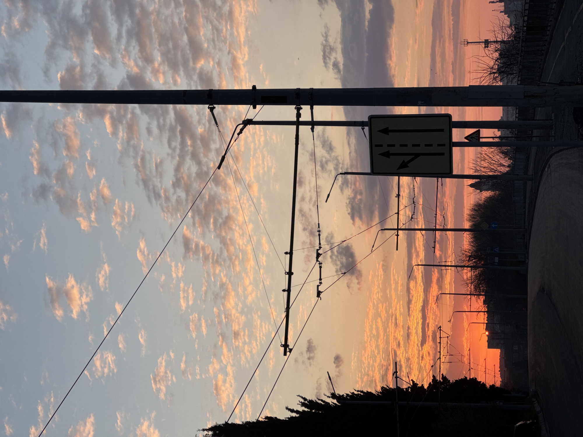 The image captures a sunset scene in Burgas, Bulgaria. The sky is filled with striated clouds tinged with orange, pink, and purple hues. Overhead electrical wires and poles associated with trolleybus or tram infrastructure cut across the sky. A rectangular traffic sign indicating lane usage is visible, featuring arrows showing a straight lane and a right-turn lane. In the background, the silhouette of buildings and trees along the horizon is visible, along with streetlights. A dark road edge is visible. The photograph appears to be taken facing east, towards the sunrise.