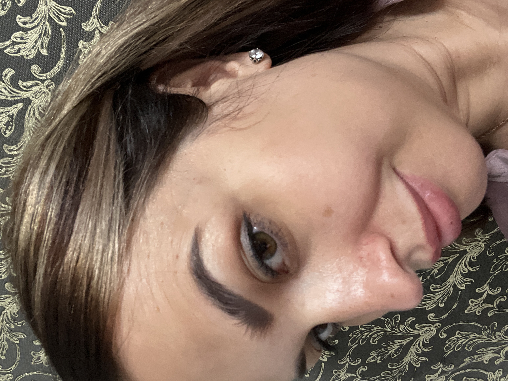 A close-up, low-angle selfie of a woman’s face is captured against a dark, patterned backdrop. The woman is looking down and to her left, with her head tilted. Her brown hair is parted and falls across her forehead and cheek. She is wearing a small, sparkling stud earring in her right ear. Her makeup includes eyeliner and a pink lipstick that appears to be slightly glossy. The lighting highlights the contours of her face, with subtle reflections visible on her skin. The background is a textured fabric with a repeating, ornate pattern in a lighter color.