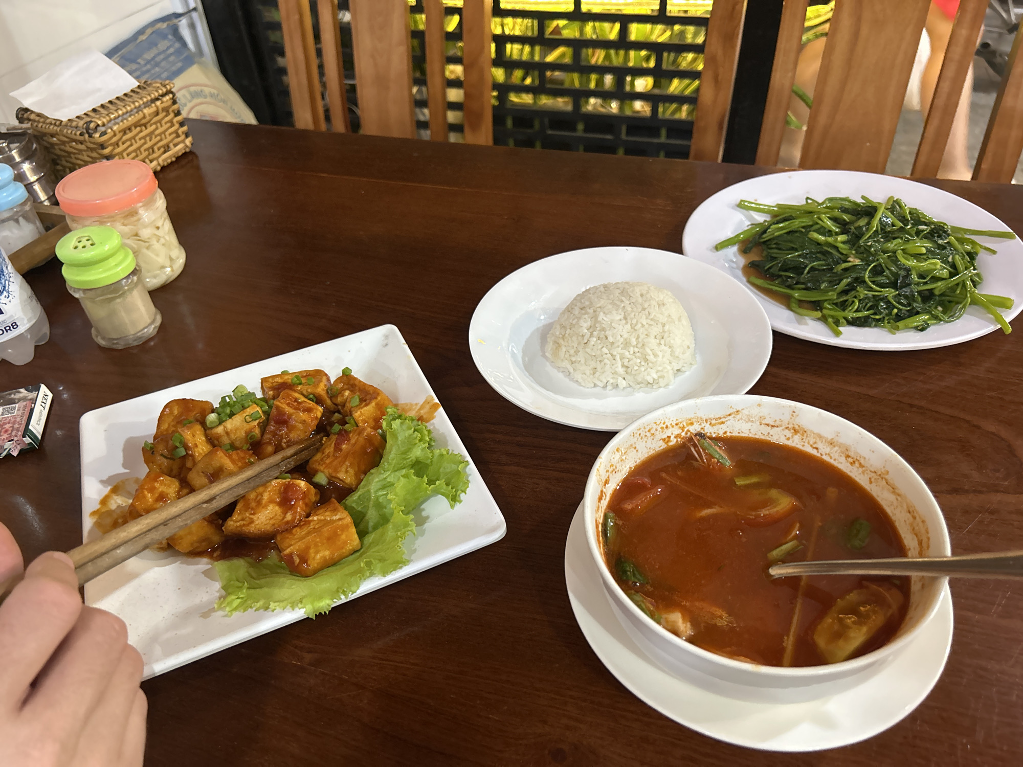 The image shows a dining table with several dishes of food, likely in a restaurant. The location is suggested to be Nha Trang, Vietnam. On the table, there is a plate of fried tofu with green onions and lettuce, a bowl of white rice, a plate of stir-fried water spinach, and a bowl of red-colored soup with a spoon inside. A person's hand is visible, holding chopsticks, and appears to be picking up a piece of tofu. Condiment jars and a tissue box are also on the table. The setting is likely indoors, with a wooden background and a window area behind the chairs.