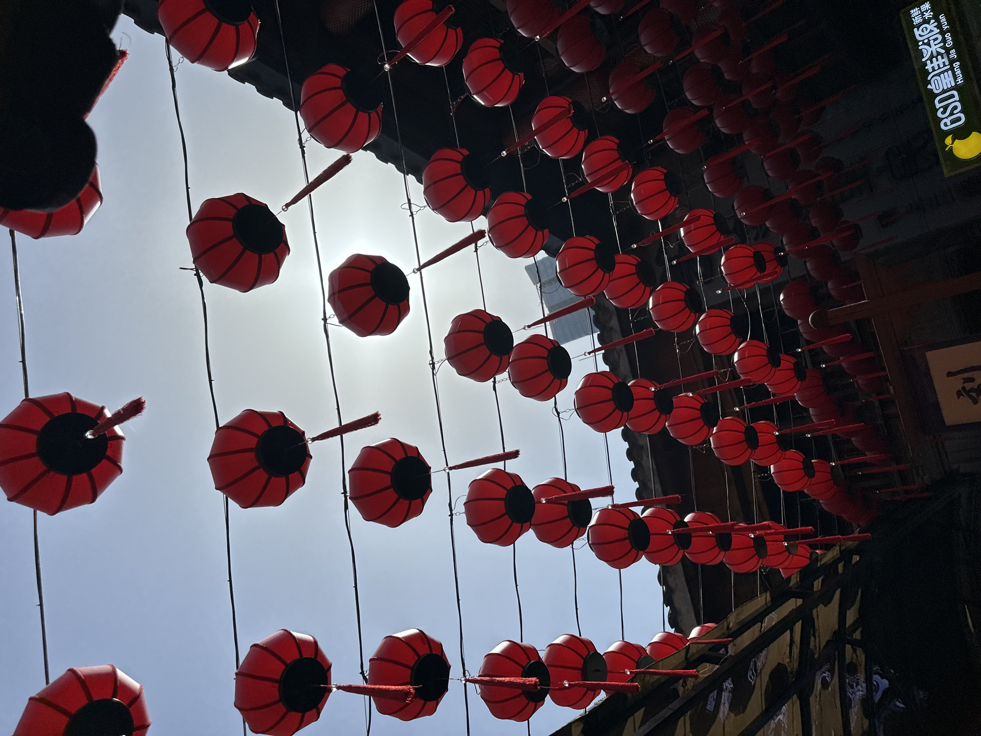 The image captures a vibrant overhead display of numerous red lanterns strung across what appears to be a street or alleyway in Yuzhong District, China. The lanterns are octagonal in shape with dark, recessed centers and feature small red tassels or decorative elements extending from their sides. They are arranged in multiple parallel rows, creating a visually striking pattern that recedes into the distance.
The photograph is taken from a low-angle perspective, looking upwards towards a bright, clear sky. The prominent sun casts a strong glare and produces some lens flare effects, indicating it is daytime, likely mid-day, under sunny weather conditions. Parts of dark, traditional-looking buildings are visible on both sides of the street, framing the overhead decorations.
No people or specific activities are depicted, but the presence of the lanterns suggests a festive, cultural, or decorative setting. On the upper right side, a sign is visible on one of the buildings. It features Chinese characters "黄家院" (Huang Jia Yuan) and English text "Huang Jia Yuan" below it, along with "GSD" and a yellow pear-shaped logo. Above "黄家院," additional Chinese text reads "巴适得很 值得体验," translating to "Very comfortable, worth experiencing." The scene conveys a sense of celebration and cultural richness.