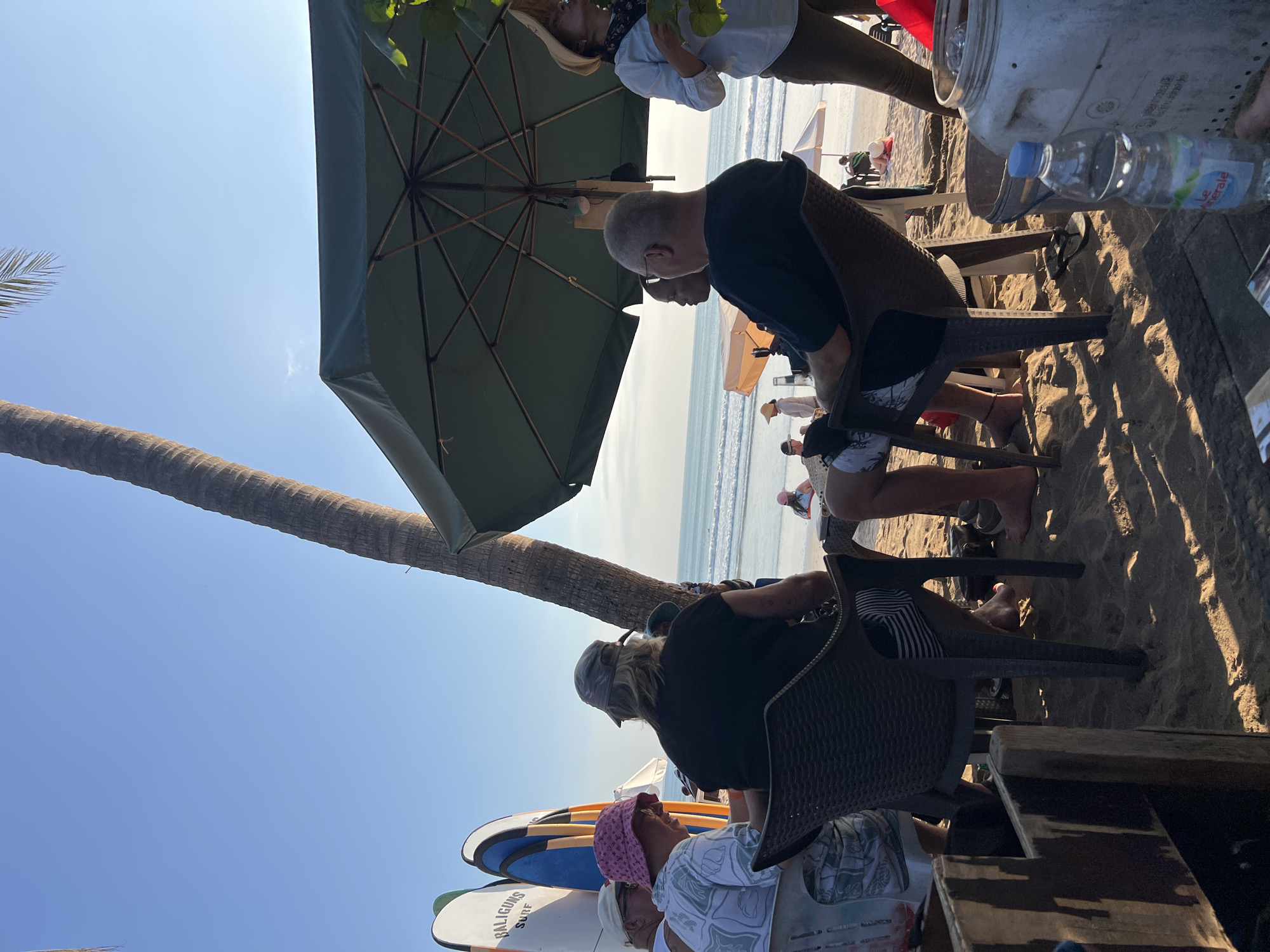 In Kuta, Indonesia, on a sandy beach, a group of people are seated at tables under a large green umbrella. Several surfboards, labeled "BALIGUNA SURF," stand upright nearby. The ocean stretches out in the background under a clear blue sky. The people are casually dressed, with some wearing hats and others exposed to the sun. The scene appears to be set at a beachside cafe or restaurant, with patrons enjoying a relaxed atmosphere. The lighting suggests it is either early morning or late afternoon.