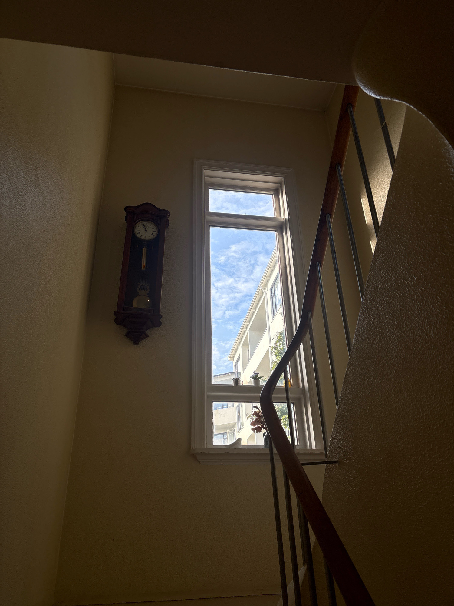The image depicts an interior view of a building's staircase. Sunlight streams in through a tall, narrow window, illuminating the textured yellow walls and a wooden banister of the staircase. On the left wall, a dark wooden grandfather clock with a pendulum hangs, showing the time as approximately 11:49. The window offers a view of another building across the way, with a bright blue sky filled with scattered clouds. Two small potted plants and a cup are visible on the windowsill. The overall impression is one of natural light in an indoor, possibly residential, setting. The specific location in Reykjavik, Iceland, cannot be determined from the visual information provided in the image.
