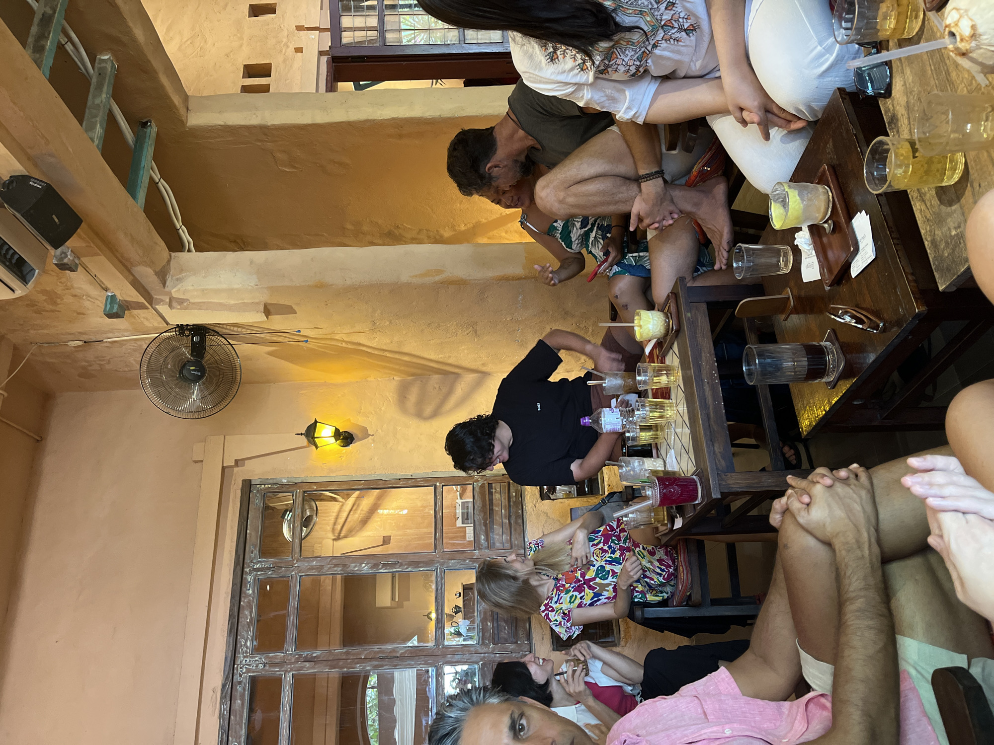 The image shows an indoor scene in what appears to be a cafe or restaurant. Several people are sitting around a wooden table filled with drinks. At least eight individuals are partially or fully visible in the shot. The table is cluttered with glasses, some containing beverages.
The environment features peach-colored walls, a large window with multiple panes, and an overhead fan. The lighting is a mix of natural light from the window and artificial light from wall-mounted lamps.
Based on the interior architecture and ambiance, the location is possibly in Đà Nẵng, Vietnam. However, the location is not definitively confirmed.
