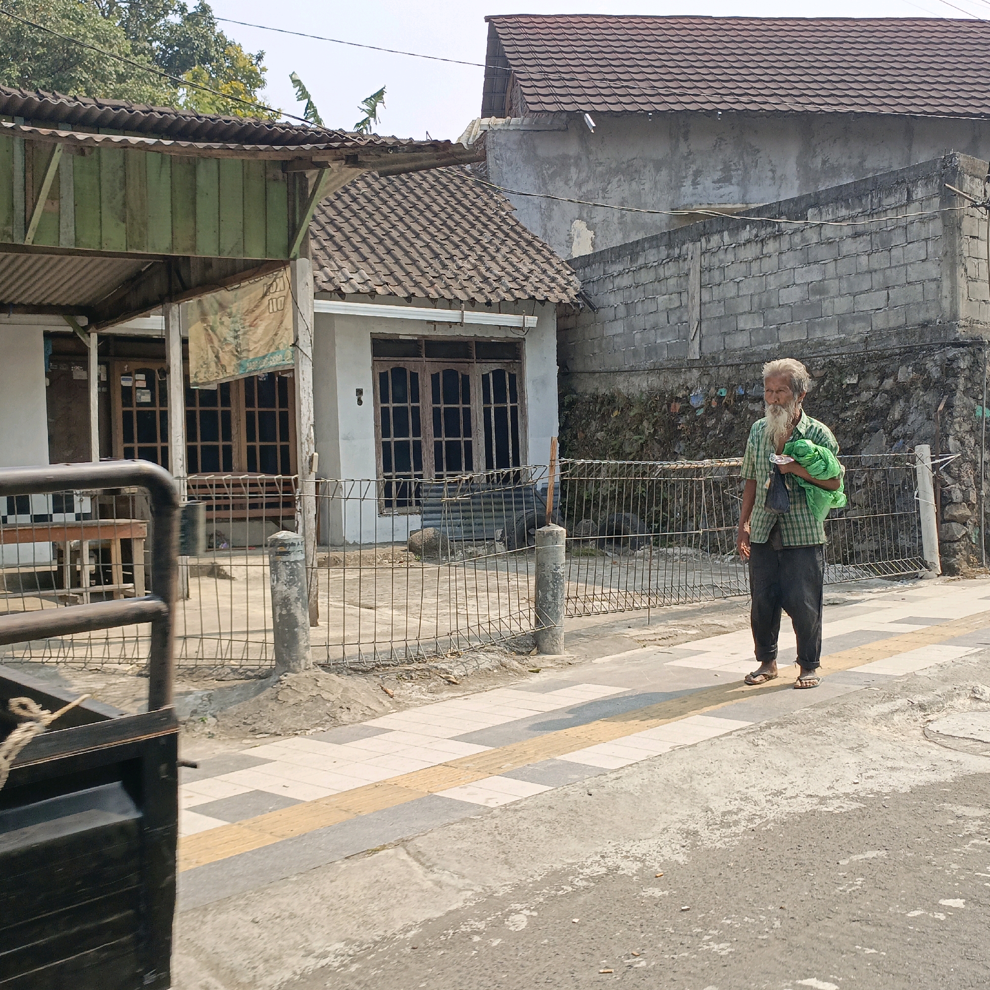 The image depicts a street scene in Salatiga, Indonesia. In the foreground, a portion of a dark-colored vehicle is visible on the left side. The middle ground features a sidewalk with a textured yellow tactile paving strip, bordering a paved road. On the sidewalk, an elderly man with a long white beard and hair is walking. He is wearing a checkered green and white short-sleeved shirt, dark pants, and flip-flops. He is carrying a bright green bag in his left arm and a dark bag in his right hand. Behind the sidewalk, a metal fence separates it from a property with a building. The building has a white facade, wooden-framed windows, and a tiled roof. To the right of this building, a higher wall constructed of grey concrete blocks is visible, with a textured stone and vegetation surface behind it. In the background, more buildings with tiled roofs and a clear sky are present. The overall impression is of a typical urban street in a warm climate.