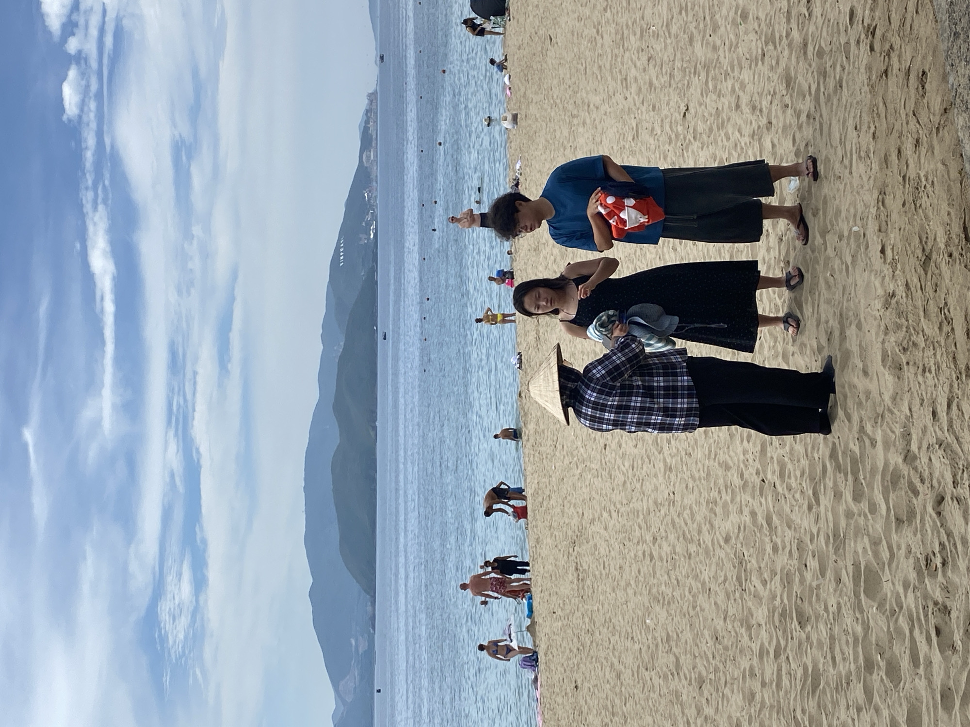 This is a photograph taken from a sandy beach in Nha Trang, Vietnam, on a partly cloudy day. The sea is calm and blue, with several people swimming or wading in the water in the background. Distant, hazy green mountains form the horizon. In the foreground, three people are standing on the sand. The person on the left wears a conical hat and a plaid shirt over dark pants. They are interacting with a woman in a black dress who is holding a towel. To the right of the woman is a young man wearing a blue t-shirt and dark shorts, holding a red and white item, possibly a child's swim float. The sand is textured with footprints and small debris. Further back on the beach, other beachgoers are visible, as are beach chairs and umbrellas. A sign with the word "VINPEARL" is visible on the mountains in the distance.