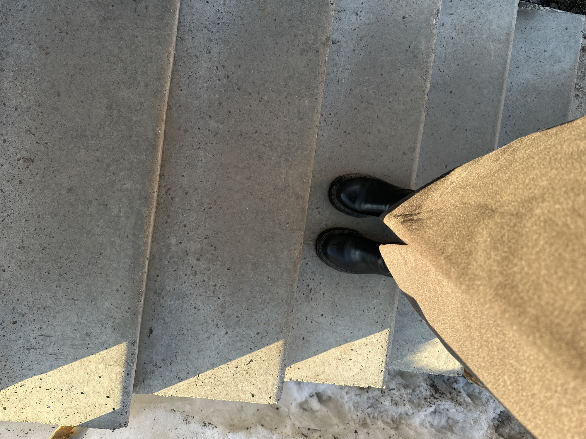 The image presents a top-down view of a person's lower body standing on a series of concrete steps in Chaikovsky, Russia. The individual is wearing black, sturdy boots with thick soles, positioned on a gray, weathered concrete step. A light brown, textured garment, possibly a coat or skirt, covers the lower right portion of the frame, receiving direct sunlight. Strong, warm sunlight casts distinct triangular shadows across the left side of the gray concrete steps, indicating a low sun angle. To the immediate left and below the steps, patches of dirty, melting snow and ice are visible on the ground, suggesting a cold weather environment. The concrete steps are unadorned and show signs of wear and minor pitting. No other people or specific city landmarks are visible.