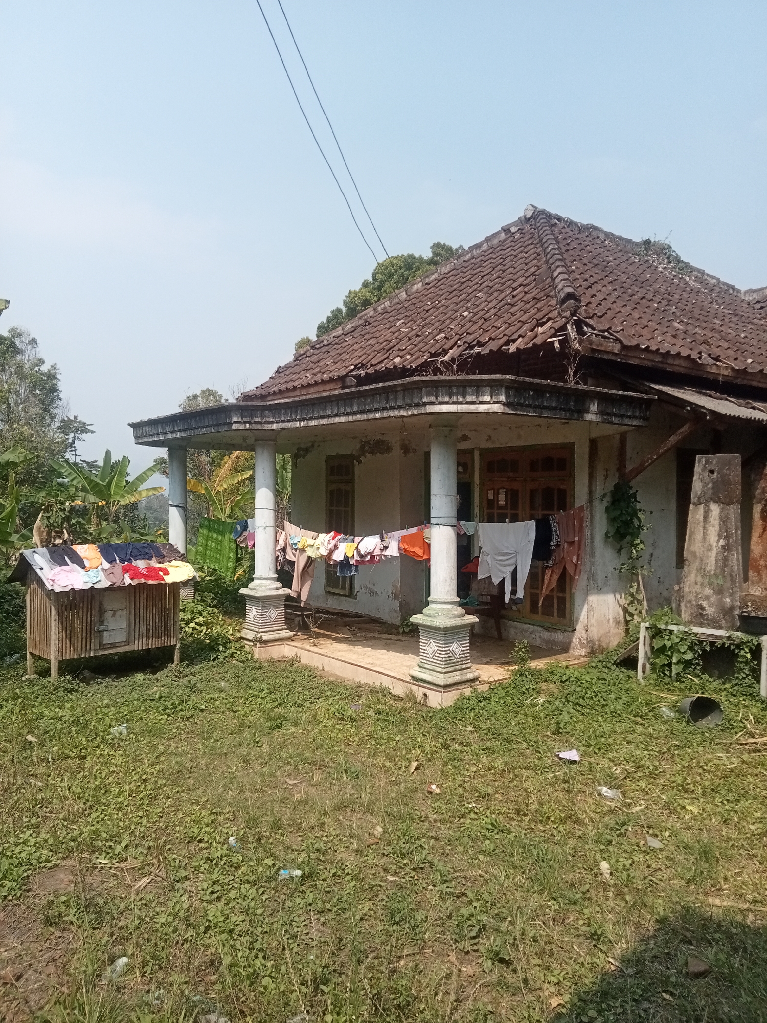 The image depicts a weathered house with a tiled roof, partially obscured by greenery. The house has a covered porch supported by columns, and laundry is drying on a clothesline in front of it. To the left, a small wooden structure with a slanted roof is also used for drying clothes, displaying a variety of colorful garments. The surrounding environment is overgrown with grass and vegetation, with some scattered plastic waste visible on the ground. In the background, banana trees and other foliage suggest a rural or semi-rural setting. The sky is clear and blue. Based on the context provided, this scene is located in Tirtomoyo, Indonesia.