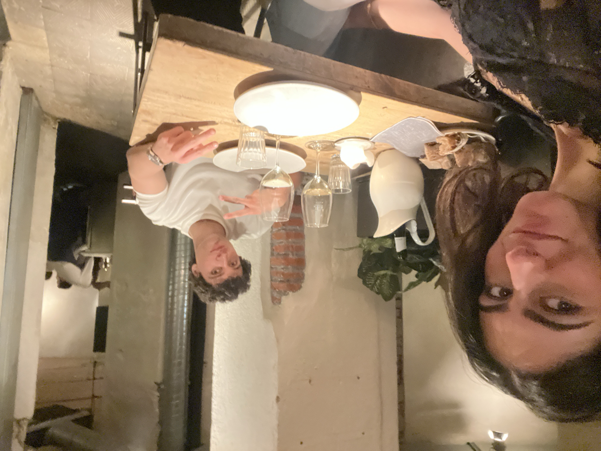 Two people are sitting at a wooden table, taking a selfie in what appears to be a restaurant. A young man in a white t-shirt is making a peace sign with his hand. The other person, a woman with long dark hair, is closer to the camera.
On the table, there are two white plates, several wine glasses, a water pitcher, a napkin, bread, and a small plant. The restaurant's walls are painted white with exposed brick. Other tables and diners can be seen in the background. The image lacks context to confirm the precise location in Madrid, Spain.
