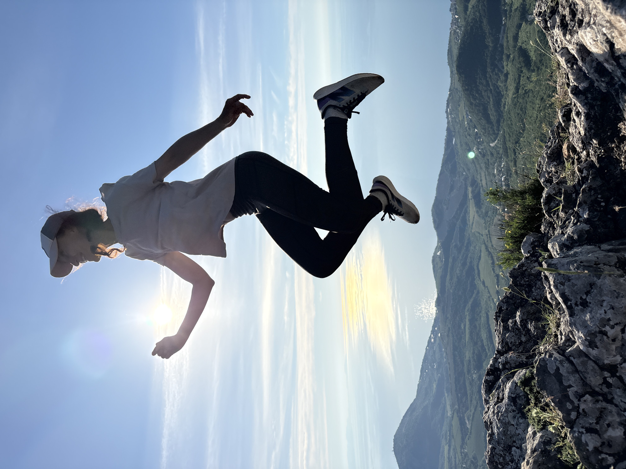 A young female, silhouetted against a bright sun, is captured mid-jump in an outdoor setting. She wears a baseball cap, a light-colored t-shirt, dark leggings, and athletic shoes. Her body is angled, with arms bent and legs lifted, conveying dynamic movement. The sun is positioned behind and to her left, creating significant lens flare and backlighting her profile, with some of her hair illuminated. The background features a vast expanse of blue sky with wispy clouds, transitioning to a distant horizon where a large body of water reflects the sunlight. Below the horizon, a panoramic view of rolling green hills and mountains with scattered buildings and roads is visible. The immediate foreground consists of rugged, grey rock formations with sparse patches of green vegetation. The scene depicts an energetic human action set against a broad, natural landscape at a high vantage point in the city of Unknown, Unknown.