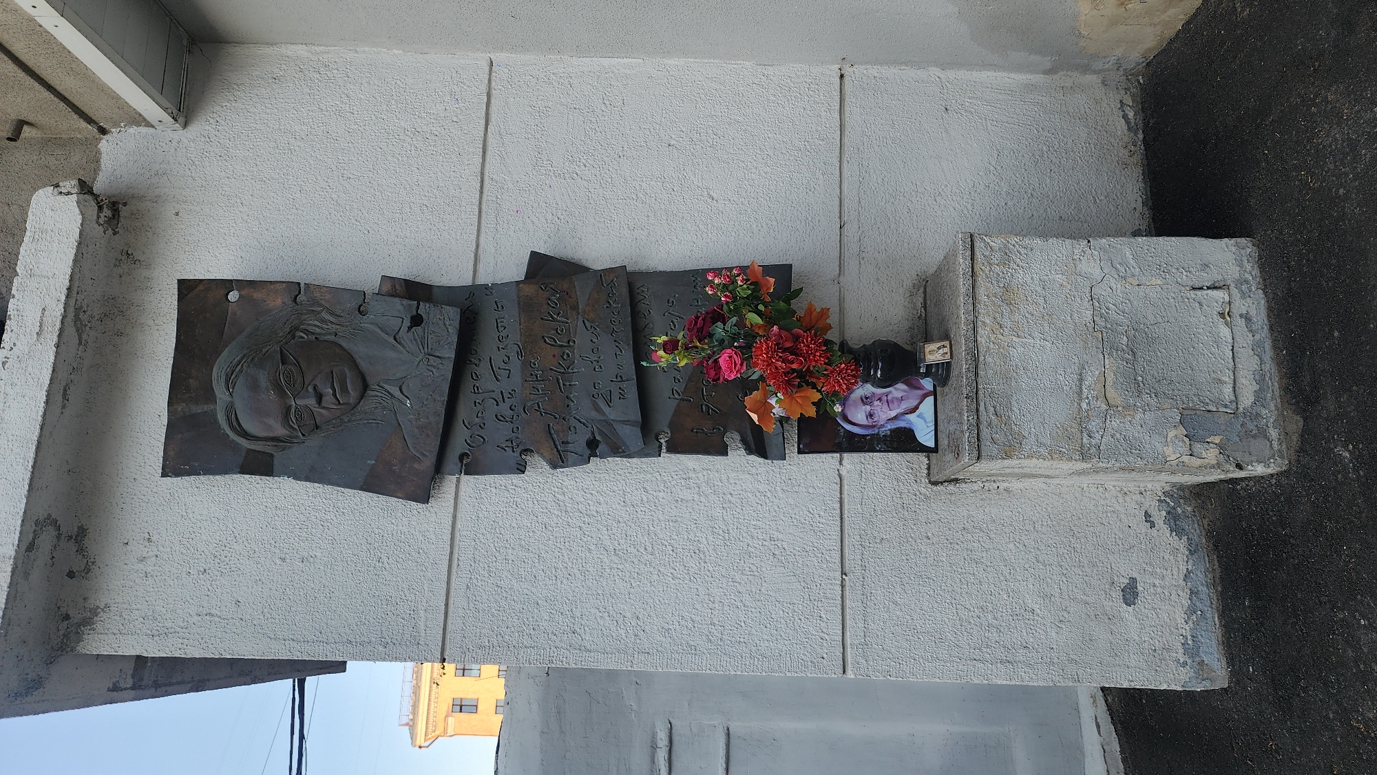 The image shows a memorial plaque dedicated to a person, possibly a writer or artist, affixed to a light-colored, textured wall. The plaque is composed of several dark metal pieces, one with a relief of a face wearing glasses, and others with inscriptions in a Cyrillic alphabet. There are flowers arranged on and next to the plaque, with a photograph of an older woman placed alongside them. The wall is adjacent to a corner where two surfaces meet, and a stone block is positioned to the right of the plaque. A building with yellow windows is visible in the bottom left corner. The scene is likely in Moscow, Russia, based on the Cyrillic inscriptions, the building's architectural style, and the overall urban environment. The photograph appears to be a modern addition to the memorial.