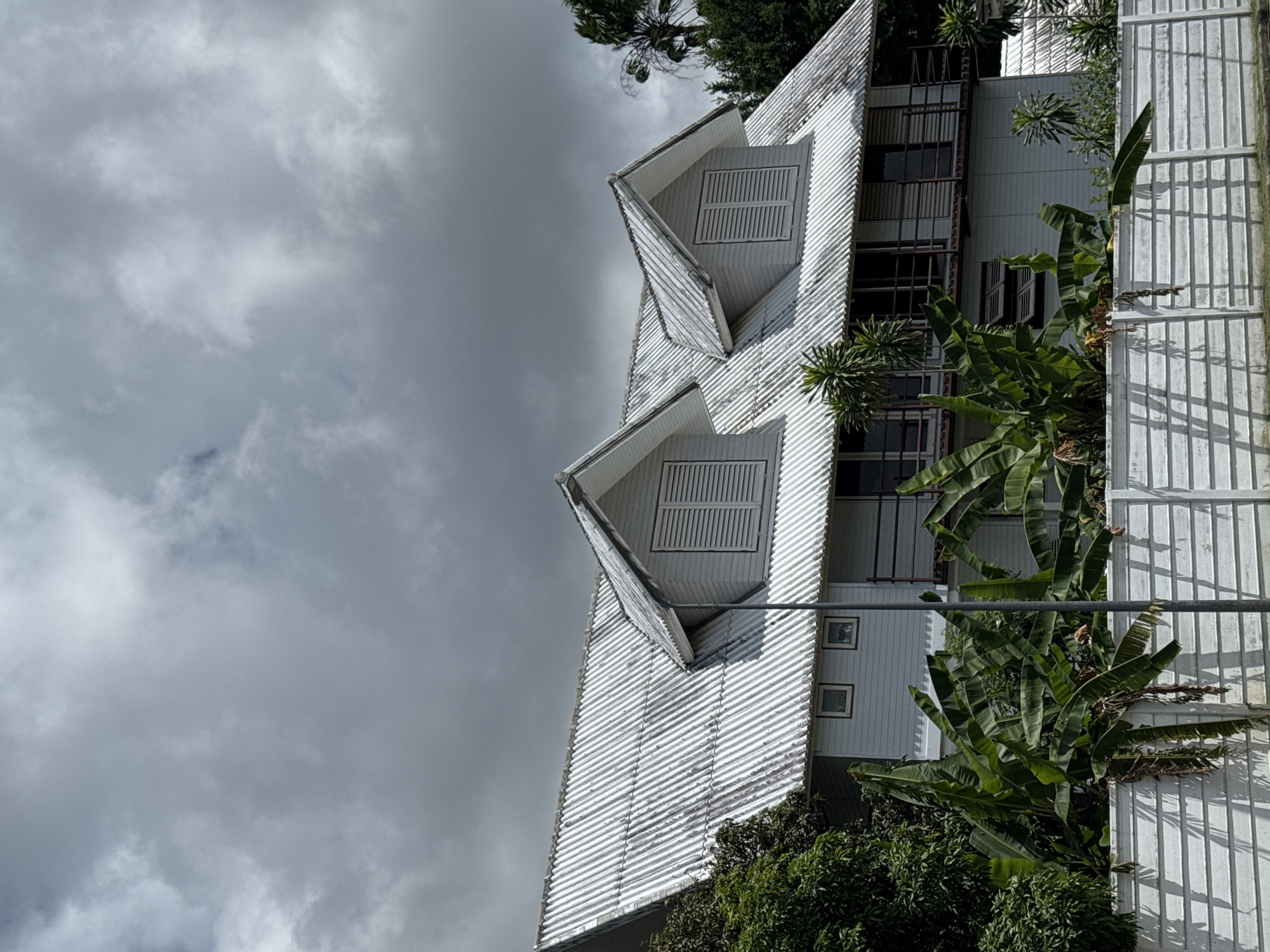 An exterior, medium shot captures a two-story, light-colored building with a corrugated metal roof. The building features multiple dormer windows with louvered shutters and balconies with railing. Lush green banana trees and other foliage partially obscure the lower portion of the building. A white concrete fence runs along the foreground, casting shadows from the vegetation. The sky above is overcast with heavy, grey clouds. The scene is set in the city of Chalong, Thailand. There are no people or animals visible in the frame, and no discernible actions or interactions are taking place.