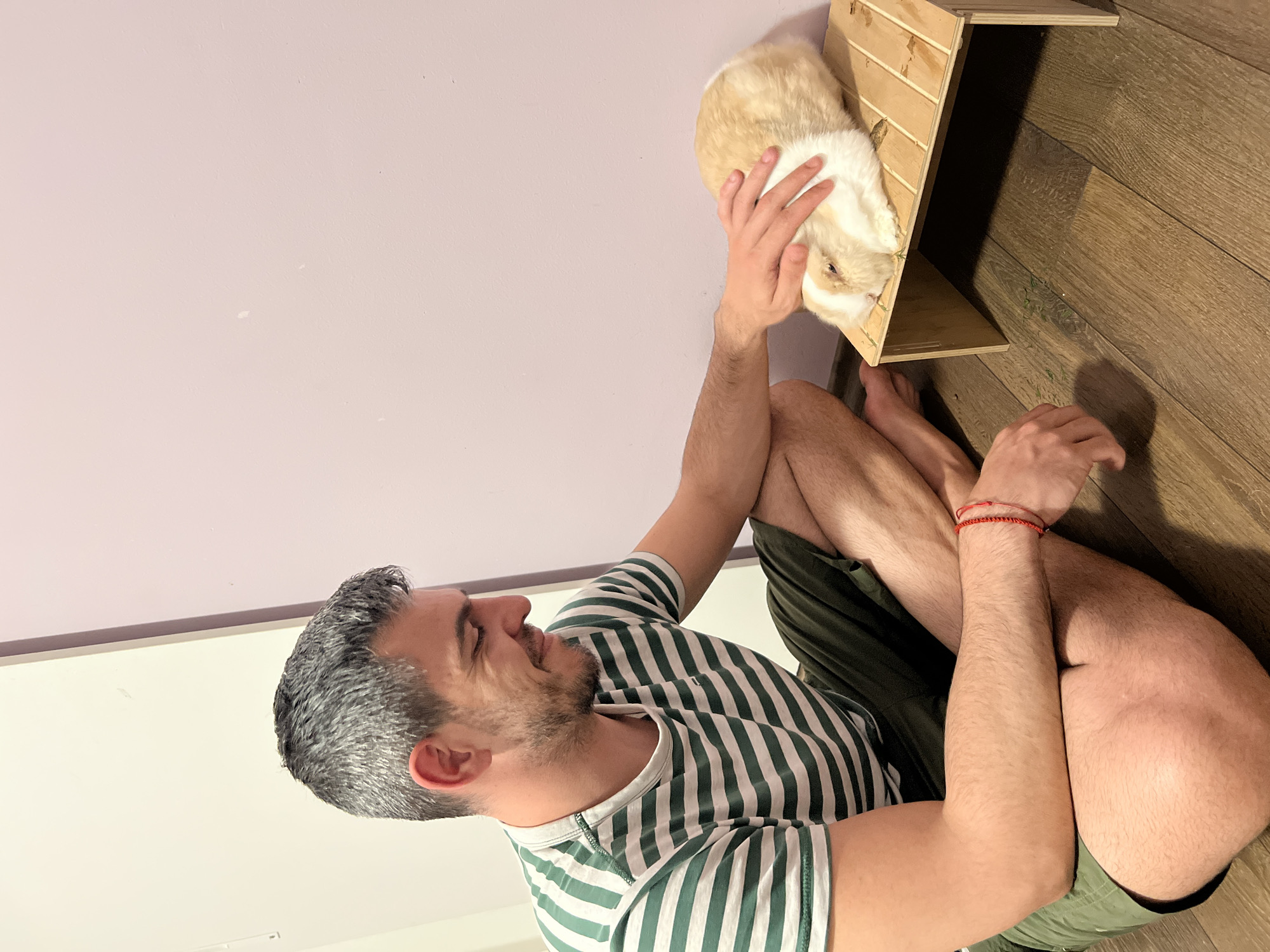 A man with salt-and-pepper hair and a beard sits cross-legged on a wooden floor. He is wearing a green and white striped t-shirt and olive green shorts. His right hand is gently petting a light brown and white rabbit. The rabbit is lying on a small, light-colored wooden platform. The background consists of a pale lavender wall on the right and a white wall or door on the left. The scene appears to be indoors, possibly in a residential setting in Sofia, Bulgaria.