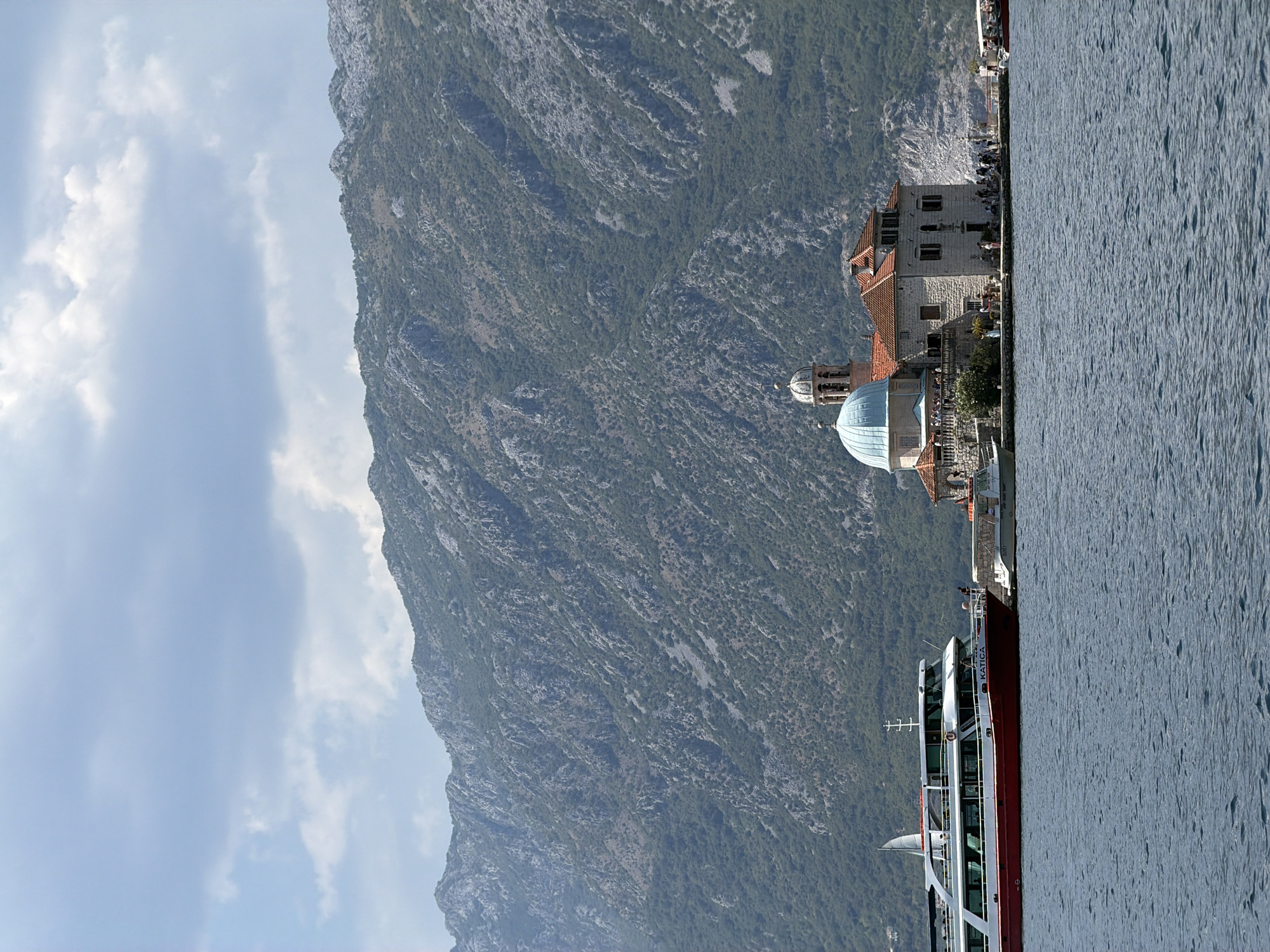 The image captures a view of the Bay of Kotor, with a prominent island church in the foreground. The island, Our Lady of the Rocks, is situated in the town of Perast, Montenegro. The church features a distinctive blue dome and a bell tower, constructed from stone. Several people are visible on the island, some appearing to be tourists.
To the left of the island, a large red and white passenger ferry, identified by the name "KATICA" on its side, is docked. Another smaller boat is visible alongside the island church. In the background, a rugged, green-covered mountain range dominates the skyline, with a partly cloudy sky above. The water in the bay appears calm and blue. The scene suggests a sunny day, typical of a tourist destination.