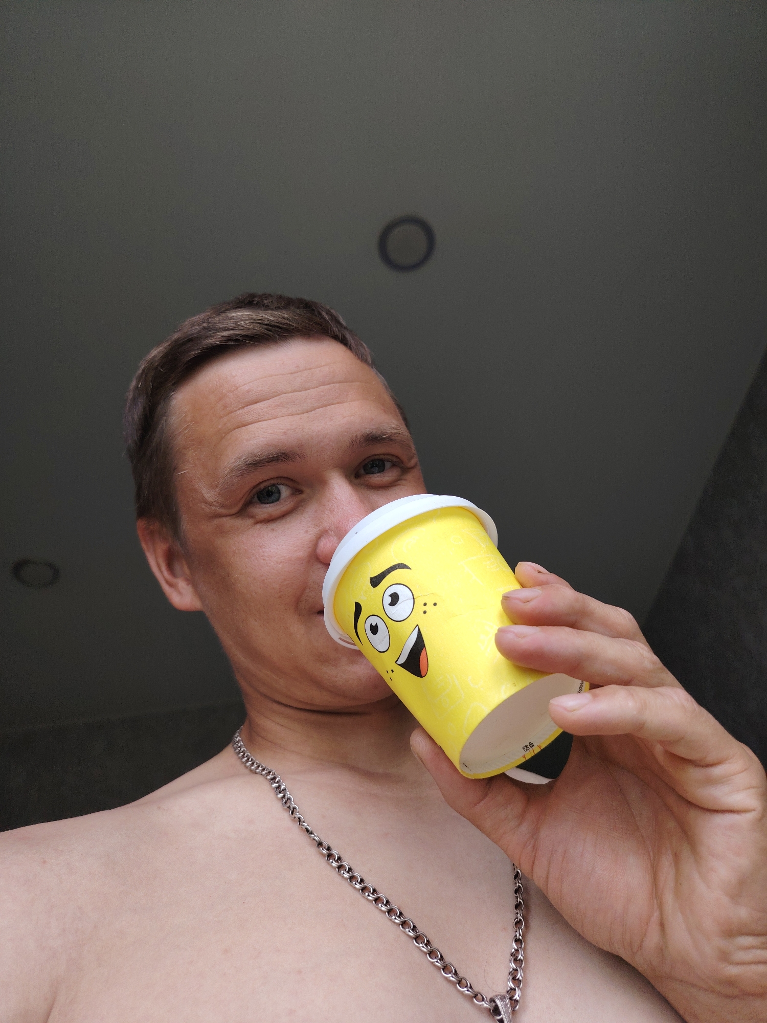 A person is shown from the chest up, holding a yellow paper cup with a cartoon face on it to their lips. The person has short, brown hair, blue eyes, and is wearing a silver chain. The background is a dark, textured wall with two ceiling lights visible. The image appears to be taken indoors. The location is not specified in the image.