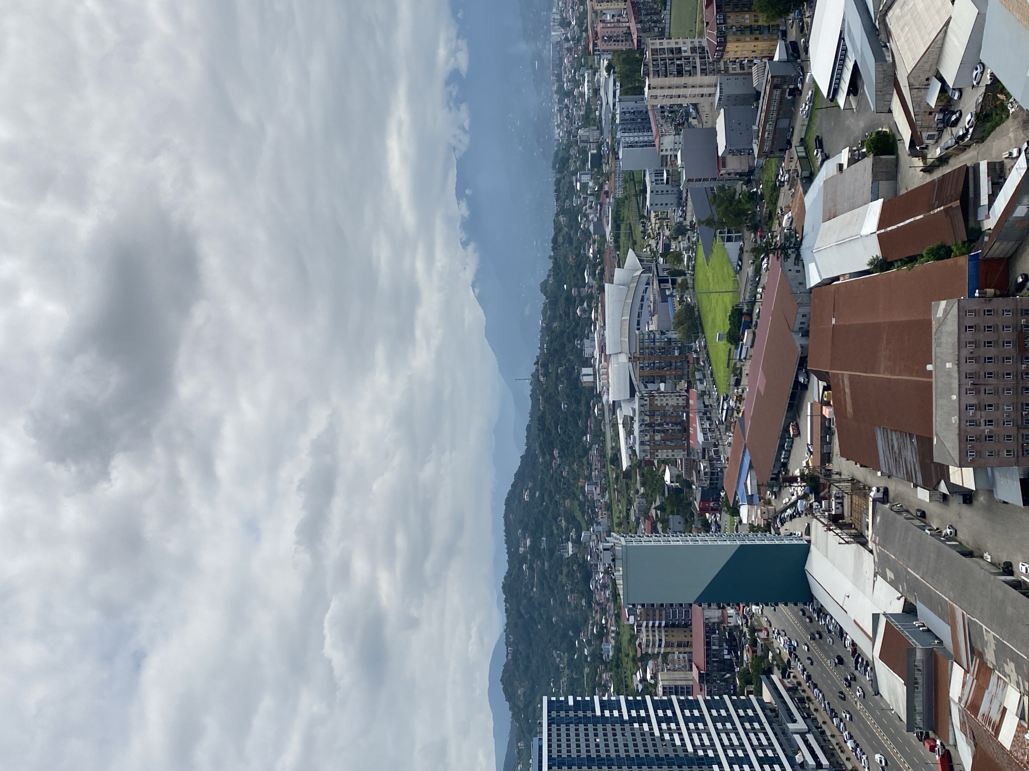 The image shows a panoramic view of a city with mountains in the background. The city appears to be Batumi, Georgia, given the presence of modern high-rise buildings alongside older structures, a stadium, and a coastal landscape visible in the distance. The sky is overcast with clouds.
In the foreground, there are several multi-story buildings, including a prominent tall, narrow building with a light teal facade and a dark teal top, positioned centrally. To its left, a large, dark blue and white patterned skyscraper is visible. The street in front of these buildings is lined with parked cars and ongoing traffic. Beyond these structures, the city extends outwards with a mix of residential and commercial buildings of varying heights and architectural styles.
Further into the mid-ground, a large stadium with a distinctive curved roof is present, surrounded by green fields and parking lots. The landscape then transitions into a densely vegetated, hilly area dotted with houses, leading to the backdrop of layered, mist-covered mountains that stretch across the horizon.
The overall scene suggests an urban environment with a significant natural landscape. The weather appears to be cloudy and possibly humid. There are no discernible people in the image, and the focus is on the cityscape and its surrounding environment. The image is taken from a high vantage point, offering a broad perspective of the urban sprawl and its natural surroundings.