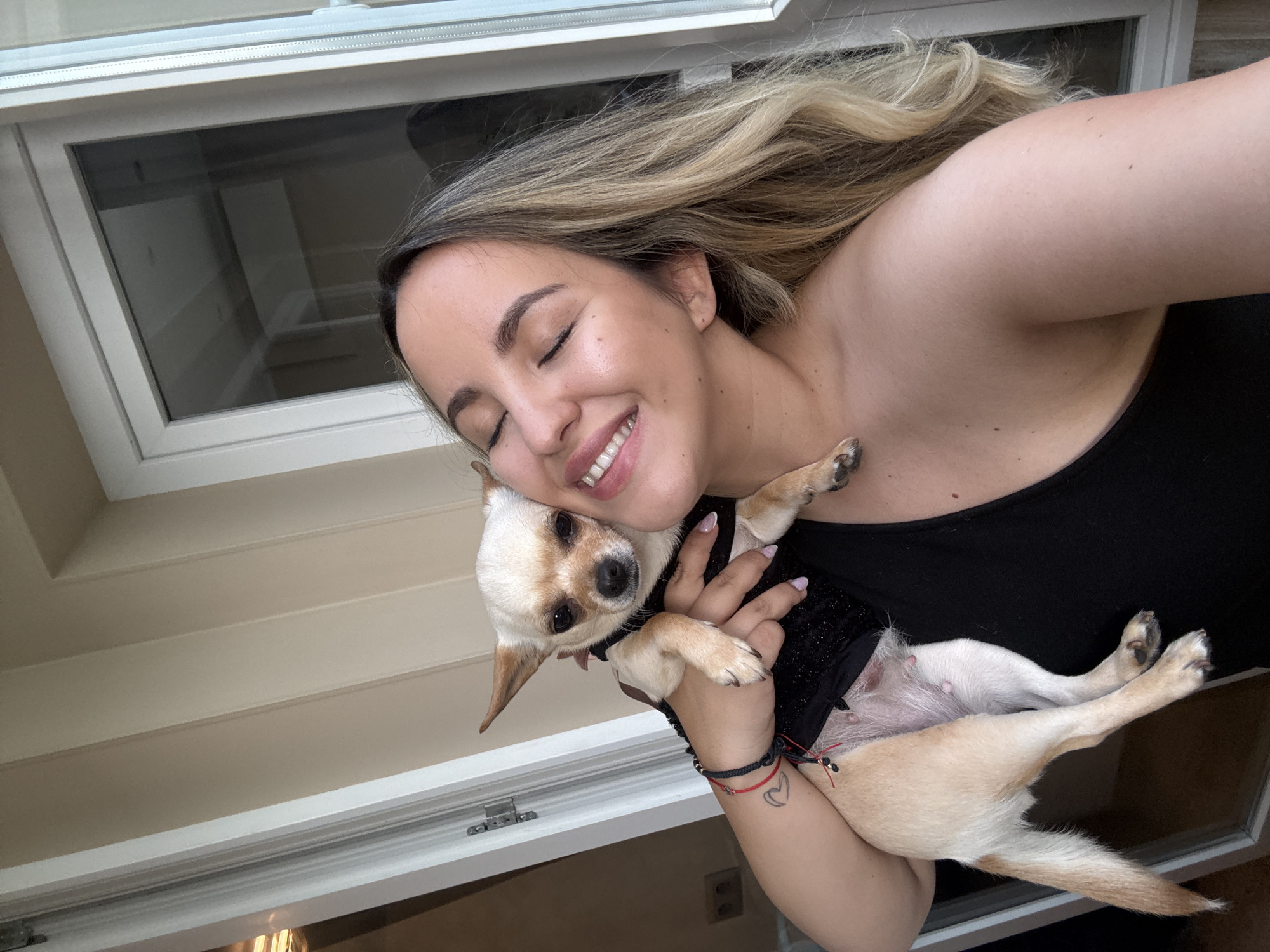 A close-up, tilted selfie captures a heartwarming moment between a woman and her small dog. The woman, with long blonde and light brown hair flowing over her shoulder, is seen from the chest up, wearing a black top. Her eyes are closed in a joyful, affectionate smile, revealing her white teeth. She is holding a small, fawn-colored dog, likely a Chihuahua, which is nestled against her neck and shoulder. The dog wears a black harness or top and looks directly at the camera with a calm expression.
The woman's left arm is visible, showing a red and a black bracelet on her wrist, along with a small, minimalist heart tattoo. The background features a white-framed window or skylight with a dark, reflective pane, suggesting an indoor setting. Below and to the side of the window, a light beige wall with horizontal paneling is visible. The overall mood of the image is one of deep affection and happiness between the woman and her pet. No visible text is present in the image.