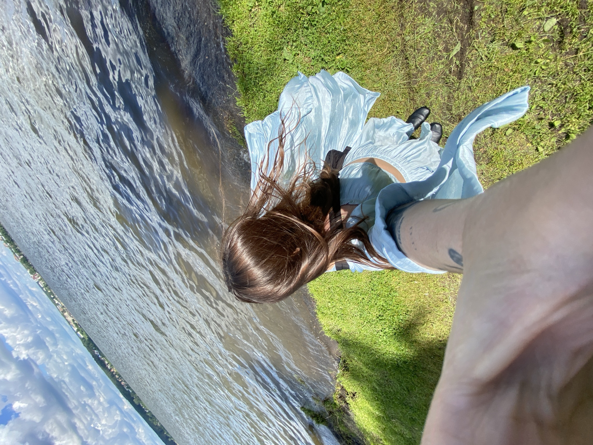 A woman in a light blue dress stands on a grassy bank next to a body of water, holding a phone to take a selfie. The water is rippling, and the sky is partly cloudy. In the distance, along the opposite shore of the water, a town is visible, which is the city of Votkinsk, Russia. The woman's hair is long and brown, and her dress is flowing in the wind. The overall scene is bright and sunny.
