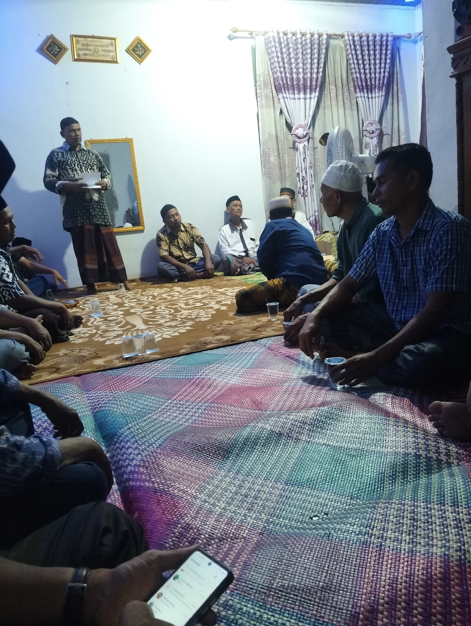 In a room with white walls, a group of men are gathered, seated on woven mats on the floor. One man, wearing a patterned shirt and a sarong, stands by a mirror and addresses the others, holding some papers. Several men are wearing peci (caps), and some are in traditional Islamic attire. In the background, curtains with floral patterns hang, and a white electric fan is visible. Several glasses are placed on a large rug with intricate designs. The scene appears to be a formal or semi-formal gathering, possibly a religious or community meeting. The lighting suggests it is indoors, potentially in the evening.