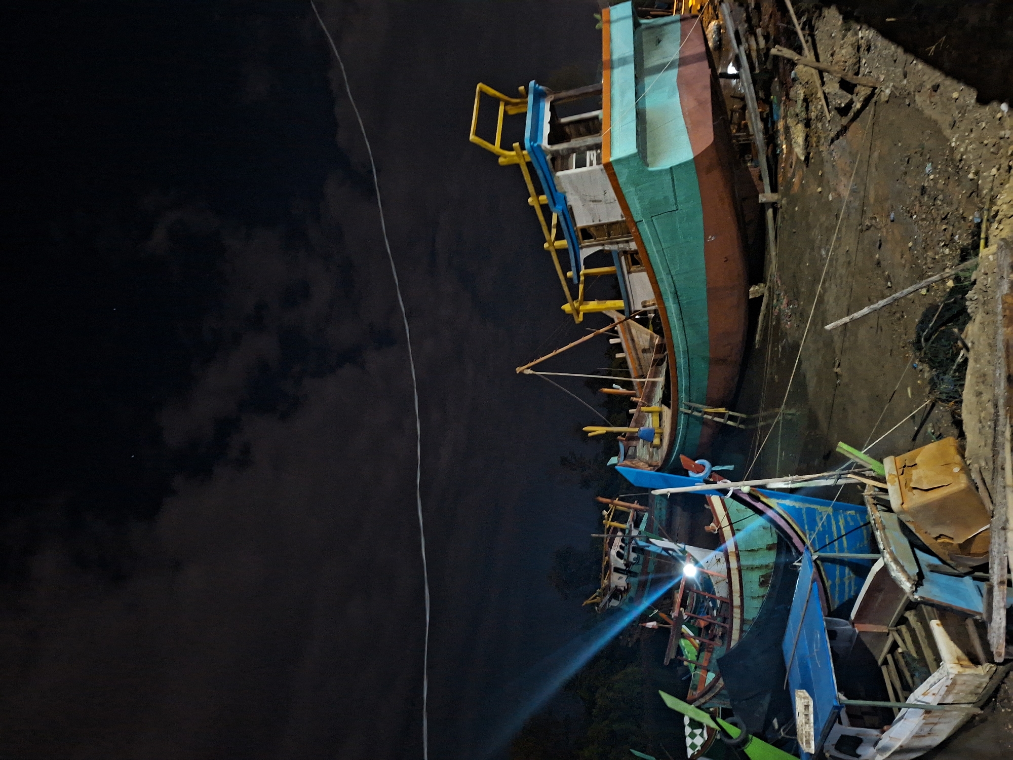 This image depicts a boatyard at night in Lhokseumawe, Indonesia. Multiple wooden boats, painted in bright blues, greens, yellows, and browns, are clustered together on land near a dirt bank. One of the boats is larger and seems more complete than the others, with visible decks and a ladder. A beam of light originates from one of the smaller boats, illuminating the immediate area. The background shows a dark sky with hints of clouds. A thin, light-colored wire stretches across the left side of the frame. The scene suggests ongoing construction or repair work on the boats.