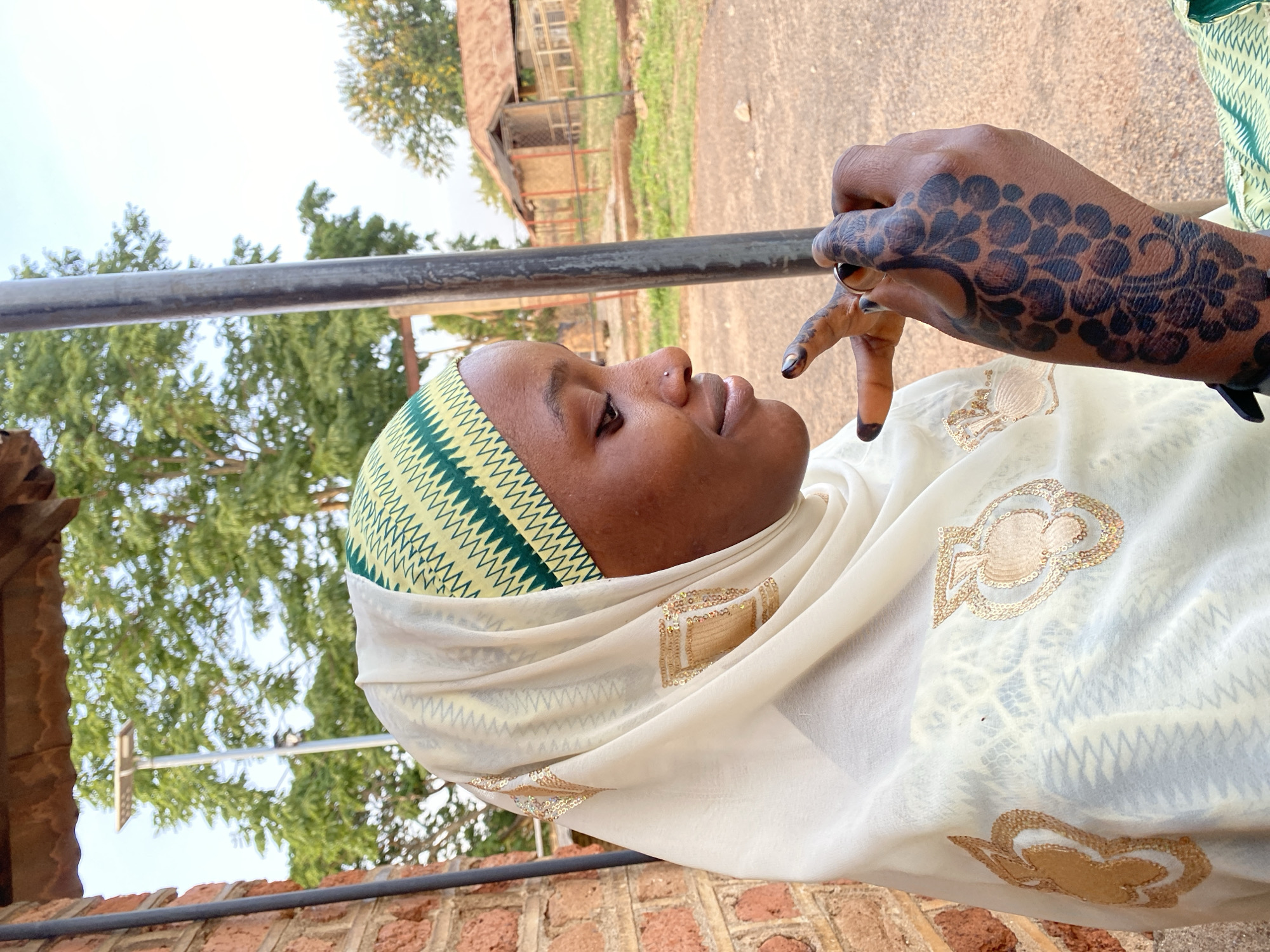 A woman with henna on her hand is visible in an outdoor setting, potentially in Vom, Nigeria, based on visual clues. She is wearing a head covering that includes a green, yellow, and white patterned headscarf along with a white hijab with a golden embellishment. Her hand, adorned with elaborate henna, is positioned near her face. The background reveals a building with a thatched roof, suggesting a rural or semi-rural location. A metal railing or bar is visible in the foreground, partially obscuring the scene. Trees and some greenery are present in the backdrop. The image appears to be shot in natural light.