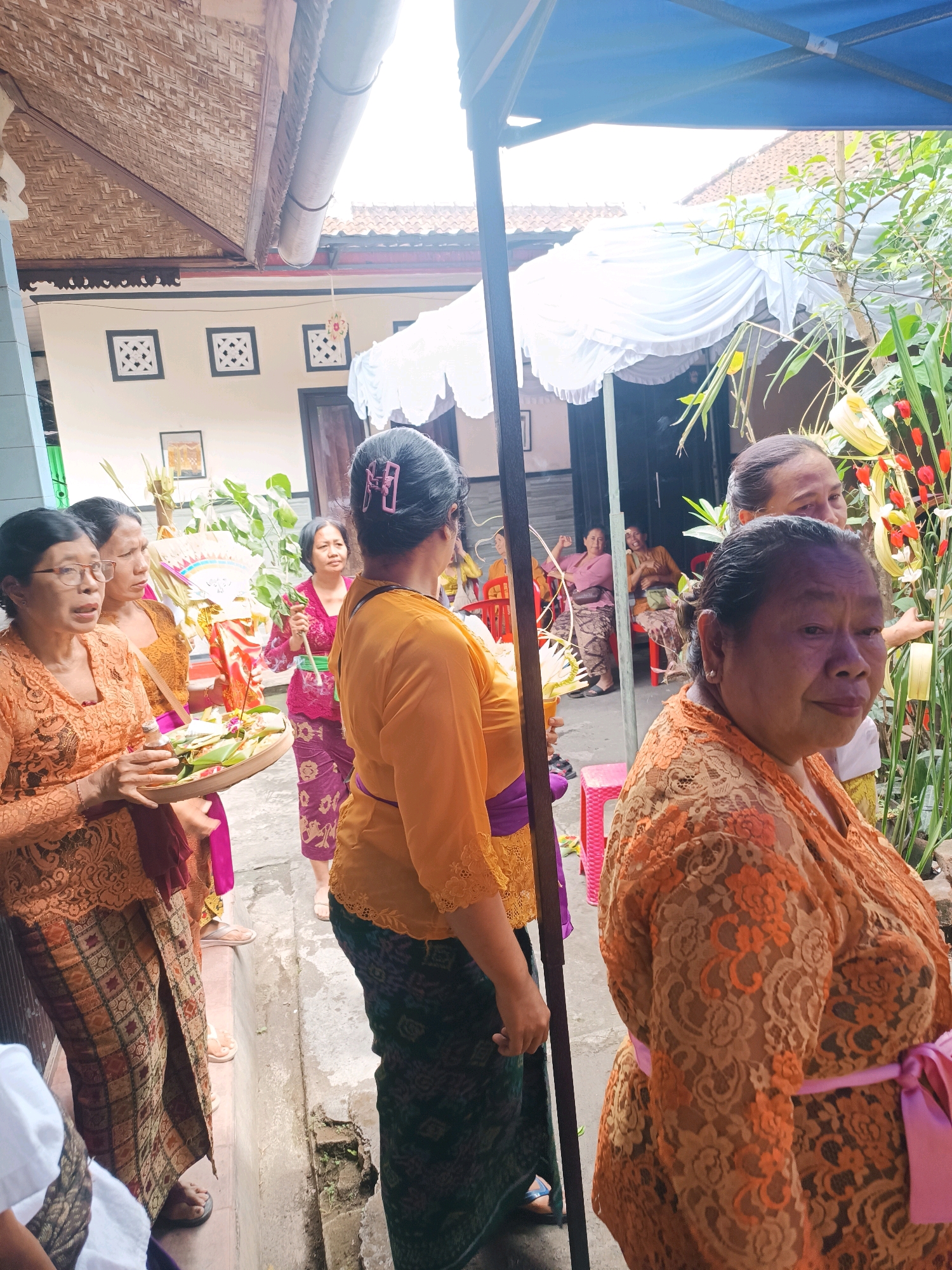 A group of Indonesian women, dressed in traditional orange and purple attire, are gathered in what appears to be a courtyard in Tabanan, Indonesia. The women are participating in a religious ceremony or cultural event. One woman in the foreground holds a woven tray filled with offerings, likely for a ritual. Other women are seen holding religious items and engaging in conversation. The background shows traditional Indonesian architecture with tiled roofs and decorative elements. A blue canopy is set up, possibly to provide shade. The overall atmosphere is one of community and spiritual observance.