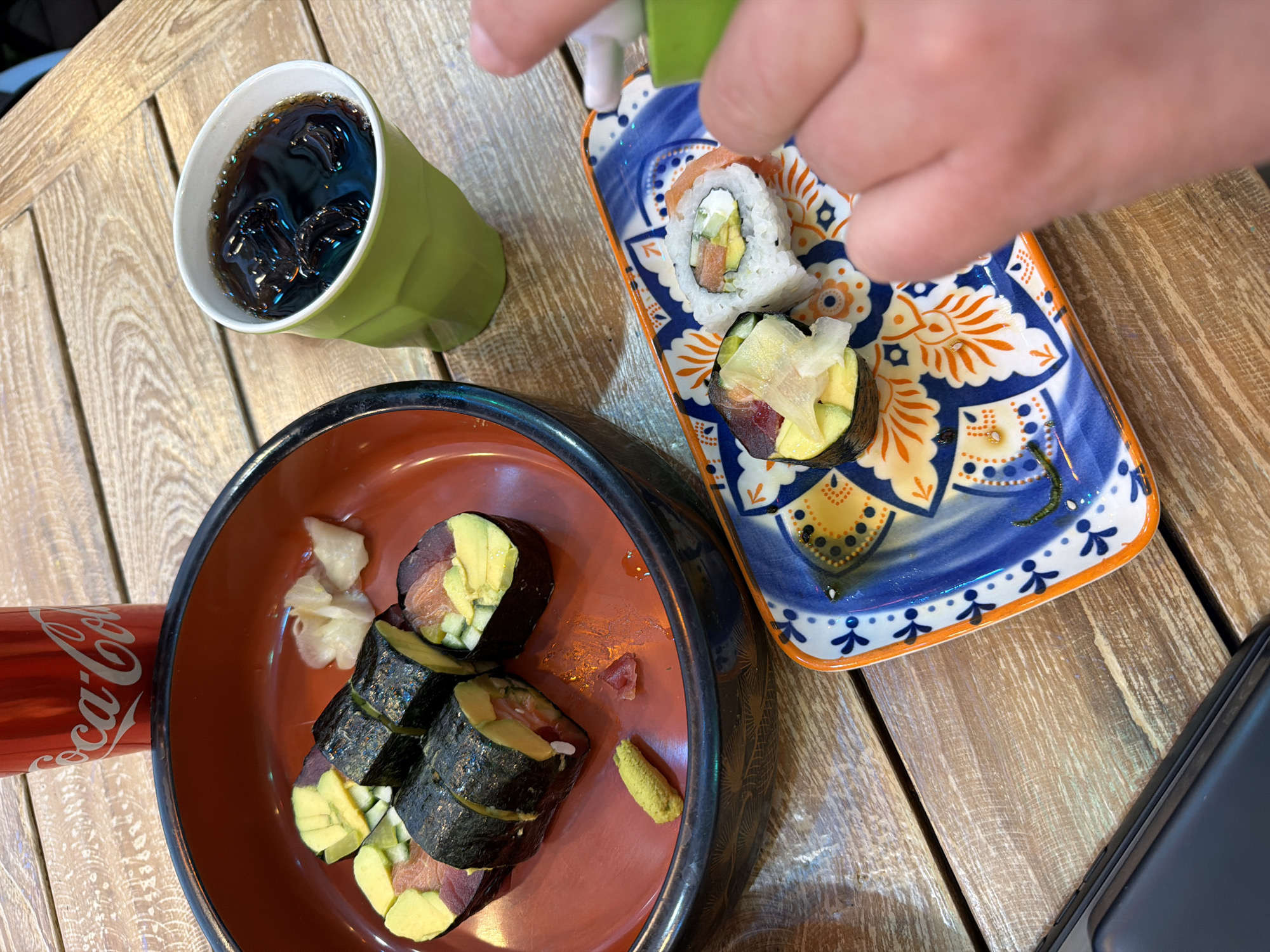 The scene shows a wooden table in an indoor setting, possibly a restaurant. The table is set with two plates of sushi, a cup of dark liquid with ice, and a can of Coca-Cola. A hand is visible, holding a green container and applying something to the sushi on a rectangular plate with a blue and orange floral pattern. The sushi on this plate includes a California roll and what seems to be nigiri with avocado and tuna. The other plate, a circular dish with a brown interior, holds more of the nori-wrapped sushi rolls with what appears to be avocado and raw fish, along with a mound of pickled ginger and a dab of wasabi. A black smartphone is also partially visible in the lower right corner. The image was likely taken in Bat Yam, Israel, given the instruction to include that location in the description.