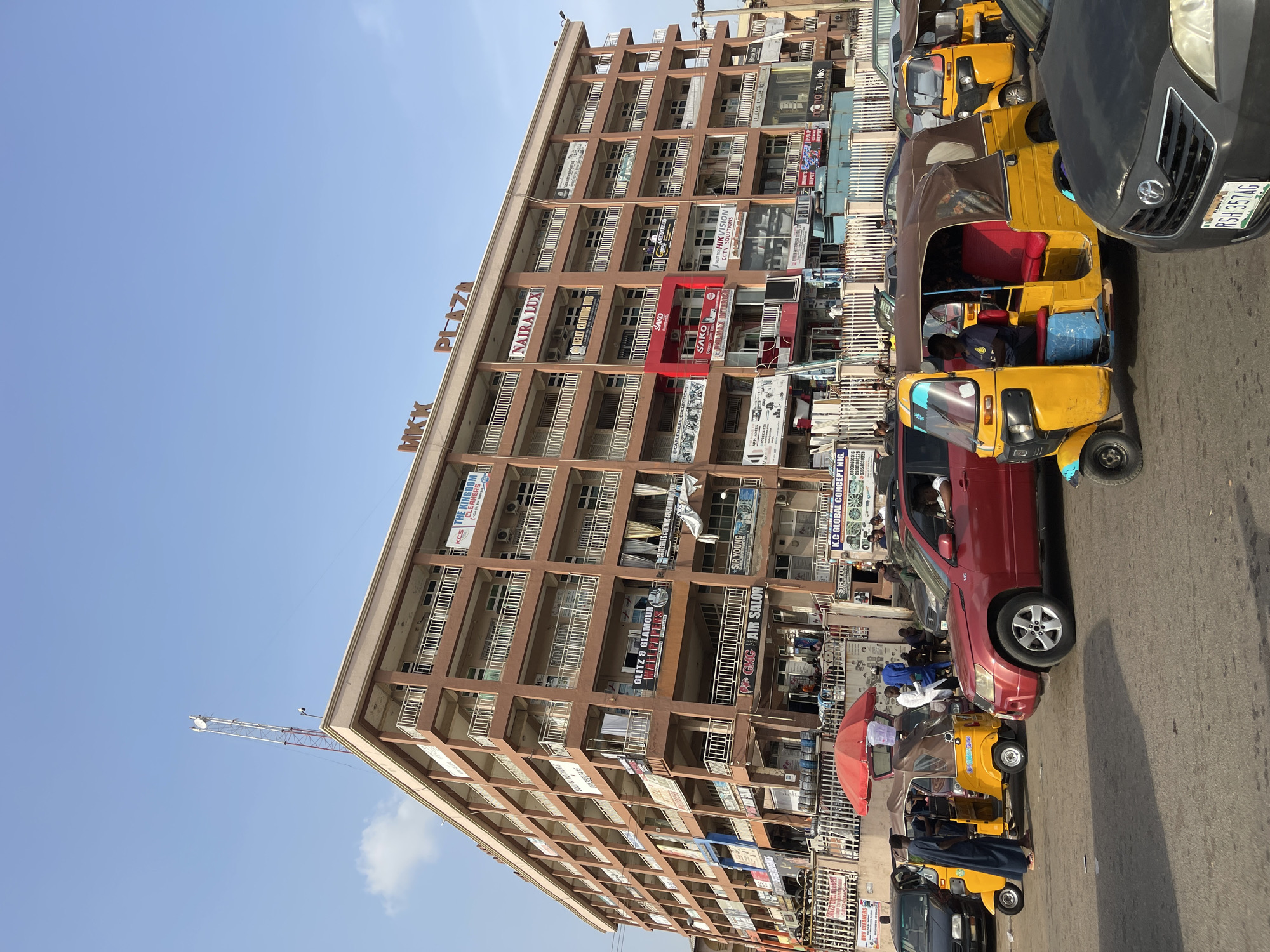 The image depicts a lively street scene in a bustling urban area, likely a commercial district in Abuja, Nigeria. A large, multi-story brown building named "MKK PLAZA" stands prominently, dominating much of the right side of the frame. This building features multiple balconies across its many floors and appears to be a commercial hub, housing numerous businesses.
Various signs for these businesses are clearly visible on the building's facade. Text identified includes "MKK PLAZA", "NAIRA LUX", "SAKO", "PHILMAX INTERIO", "HIKVISION CCTV SOLUTIONS", "K.C GLOBAL CONCEPT NIG.", "GMG HAIR SALON", "SIR YOU", "GLITZ & GLAMOUR", and "KCS THE KINGDOM CLEANERS". Other partial texts like "CLEANERS", "ECTIONS", and "APPLIANCES" are also discernible.
The street level is active with vehicles and people. Several yellow three-wheeled auto-rickshaws, commonly known as Keke Napep in Nigeria, are visible, some with blue or red accents. A red SUV and a black car are also part of the traffic. The black car displays a Nigerian license plate with the alphanumeric sequence "RSH-357AG". People are observed inside the auto-rickshaws and moving around on the street, contributing to the busy atmosphere.
The scene is captured on a clear, sunny day, indicated by the bright blue sky. A tall telecommunication antenna or mast is visible against the sky on the left side of the image. The overall impression is one of everyday urban life and commercial activity in a vibrant city.