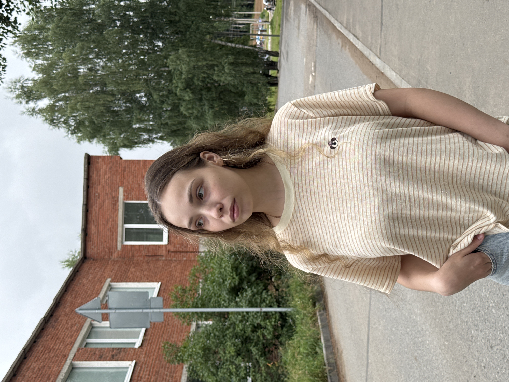 A young Caucasian woman with wavy blonde hair stands on an asphalt road in front of a brick building and a line of trees. The woman is wearing a cream-colored t-shirt with thin horizontal stripes and a small embroidered patch on the left side of her chest. She has her right hand in her pocket and is looking off to her left. The building behind her is made of red brick with white-trimmed windows. A signpost with a gray triangular sign is also visible. The area appears to be a residential or industrial area in Noviy, Russia, with overgrown vegetation along the side of the road. The sky is overcast.