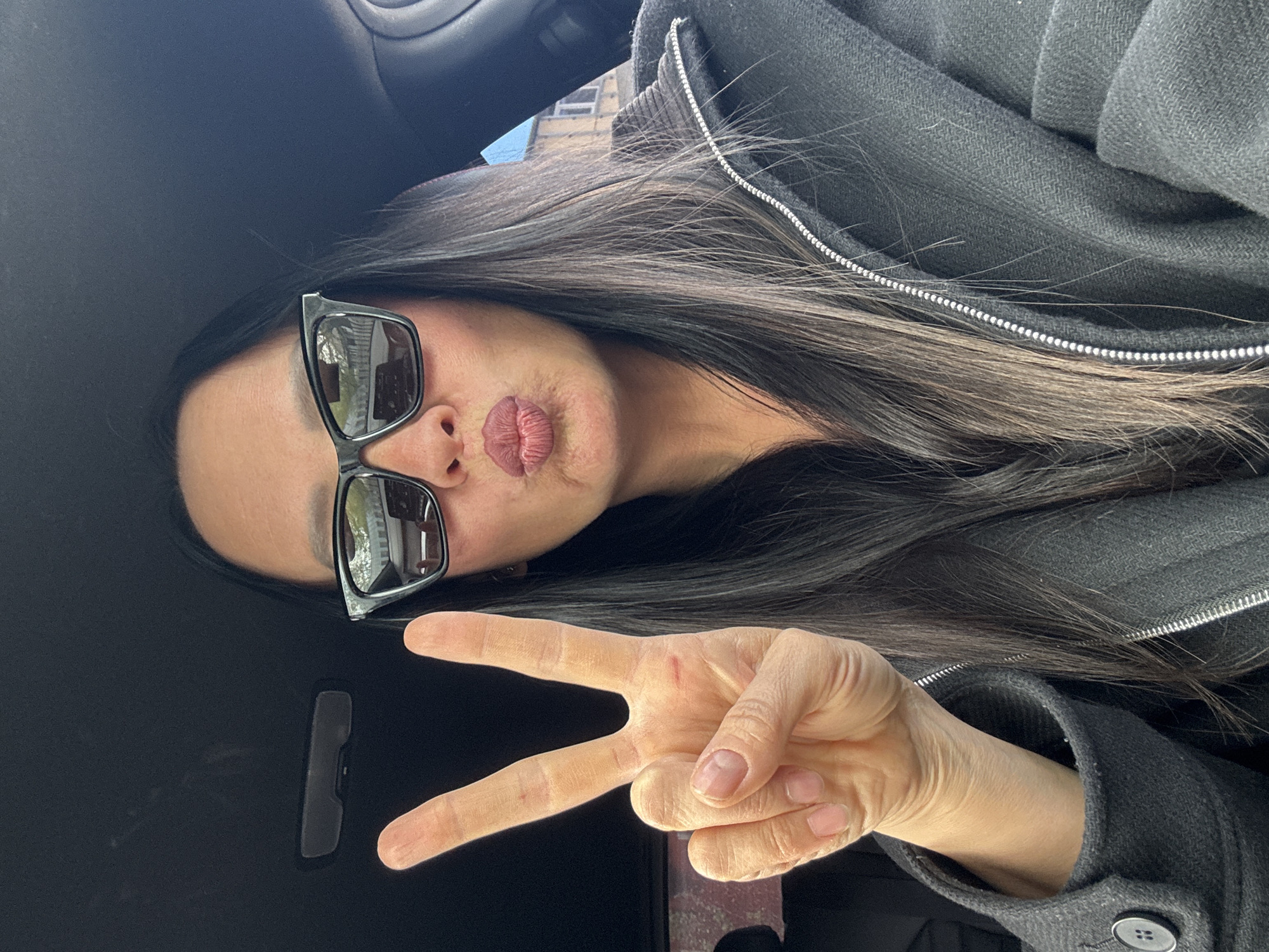 A woman with long, dark hair is pictured in what appears to be the interior of a car. She is wearing sunglasses and a dark-colored jacket with a zipper visible near her hair. She is making a "peace" sign with her hand and puckering her lips into a "duck face" pose. The background of the image shows a glimpse of a building, which is inconclusive to determine the exact location in Chaikovsky, Russia.
