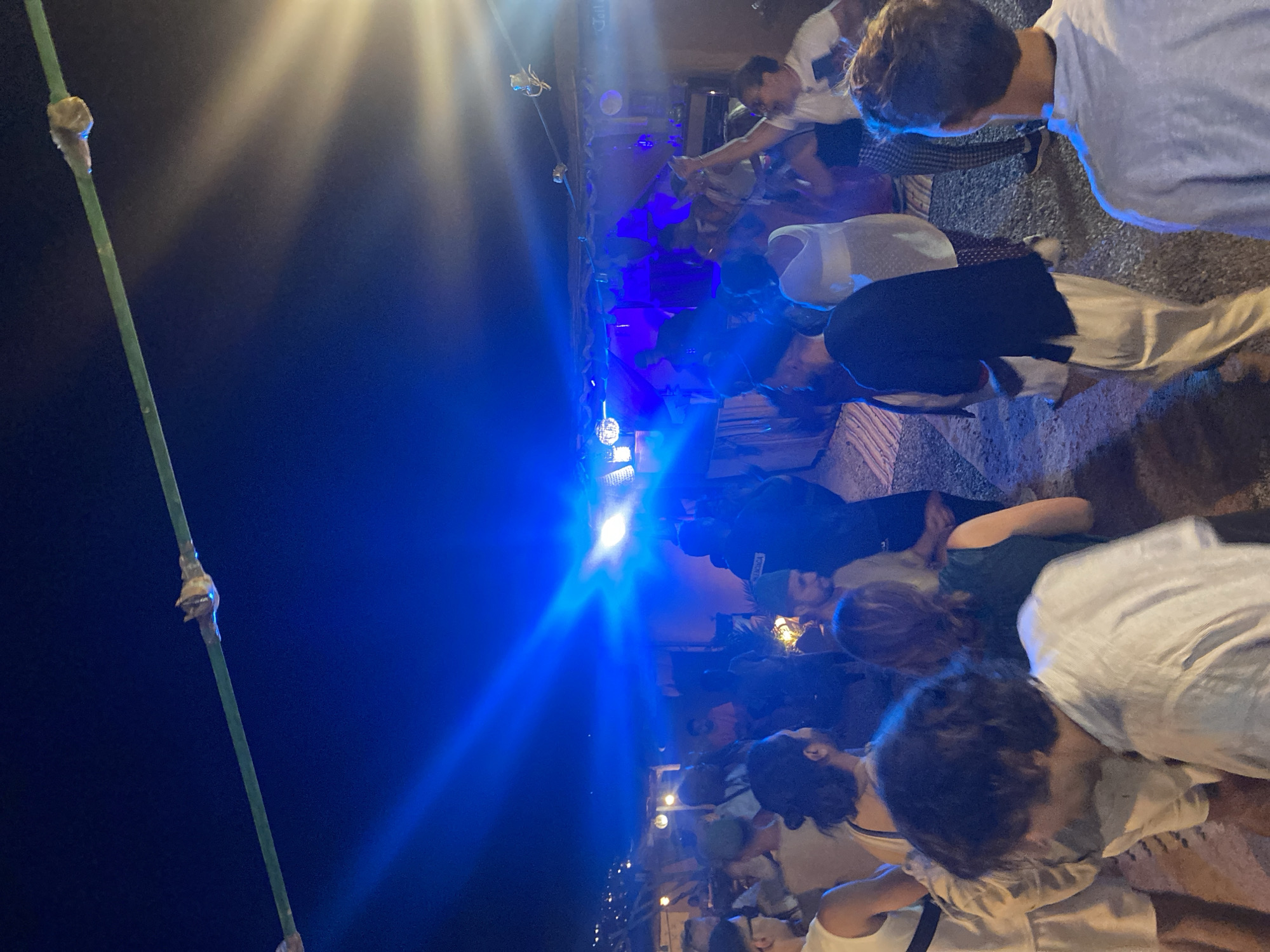 The image depicts a nighttime scene at a gathering or event in Agafay, Morocco. A crowd of approximately 15-20 individuals is visible, many of whom appear to be dancing or socializing. The lighting is predominantly blue and white, emanating from stage lights or spotlights, creating a vibrant and energetic atmosphere. Some individuals are dressed in casual attire, including shirts, shorts, and dresses. The background suggests an outdoor setting, possibly a desert camp or a venue with a rustic, open-air structure. A green cable is visible strung across the top of the frame, with small objects attached to it. The overall impression is one of a lively social event taking place after dark.