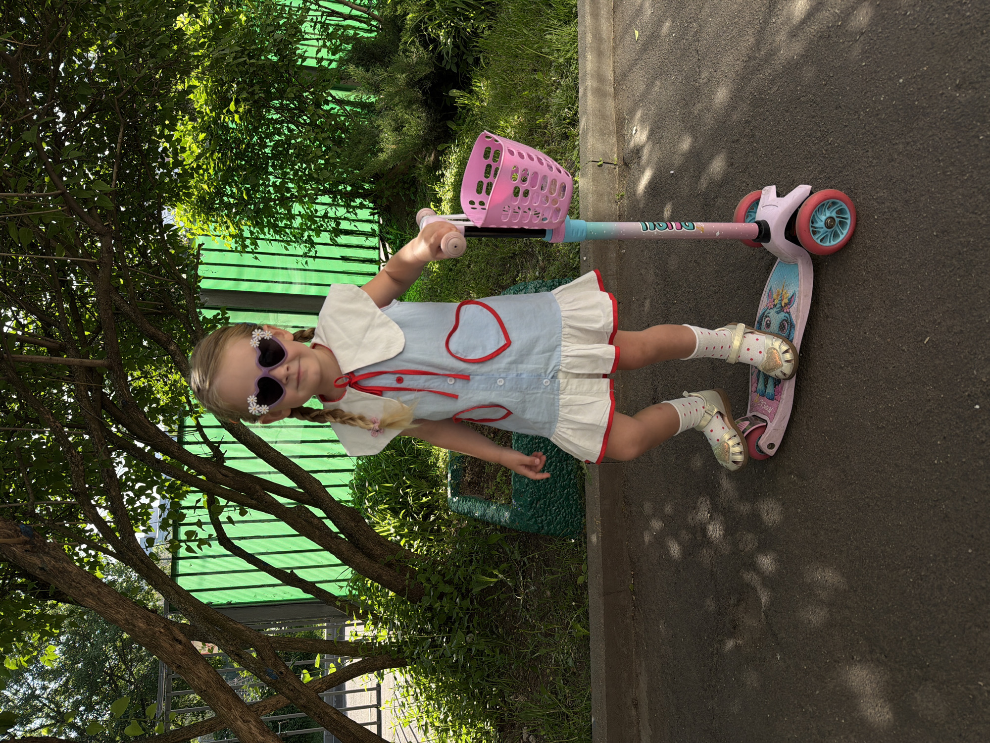 A young girl stands on a scooter with a pink basket attached, outdoors on what appears to be a paved surface near a green fence and trees. She is wearing sunglasses with a flower decoration, a light blue dress with red trim and white ruffles, and light-colored socks and sandals. Her hair is styled in braids. The background includes greenery and urban infrastructure. The precise location in Moscow, Russia is not discernible from the image.