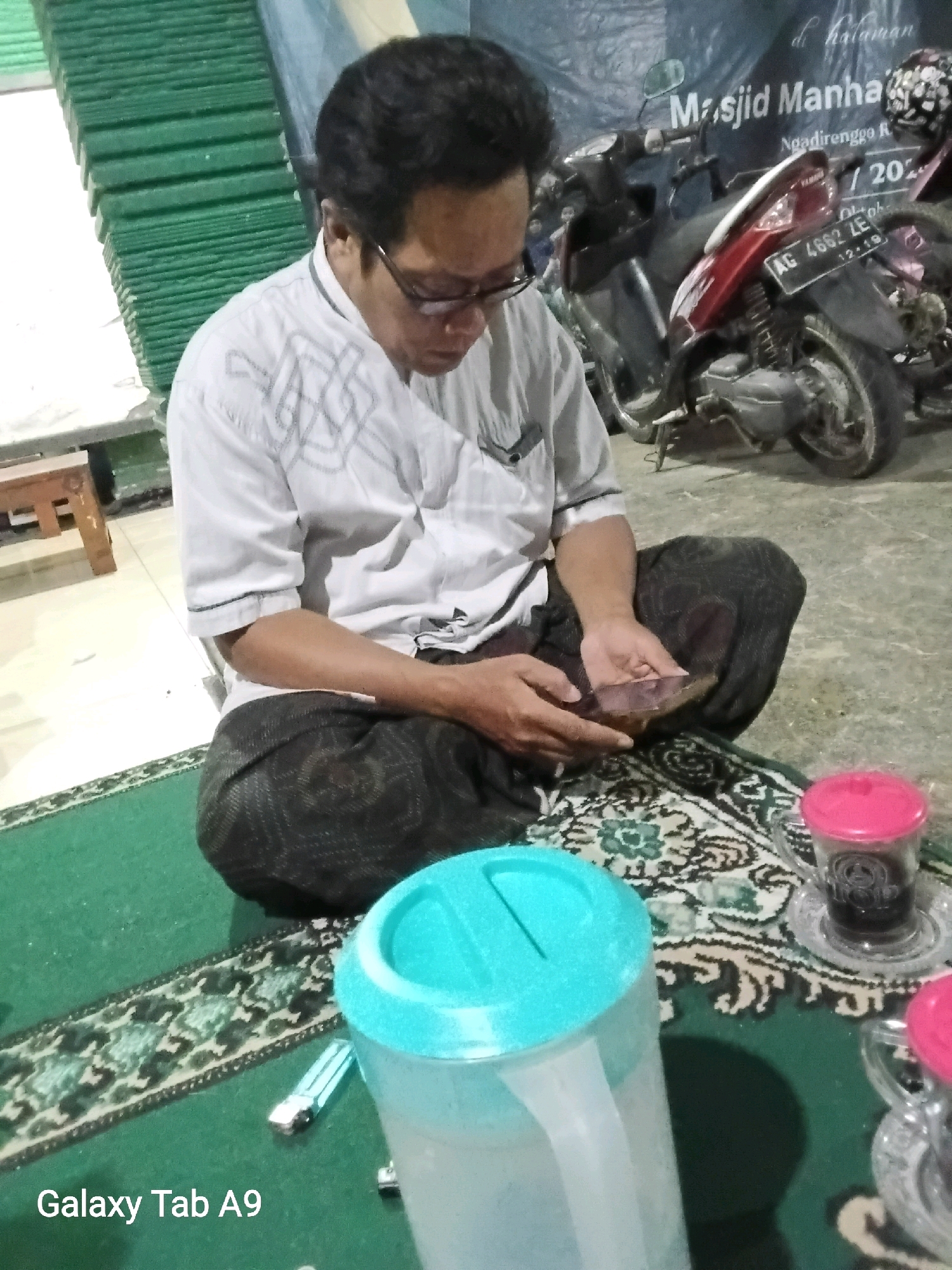A man wearing glasses and a white patterned shirt sits on a green rug in front of a motorcycle and a banner. The man is holding a smartphone in his hands. There are two cups with pink lids and a pitcher with a blue lid on the rug. The banner behind him reads "Masjid Manahan Ngadirenggo Re 2023". This suggests the location is Masjid Manahan in Ngadirenggo, Trenggalek, Indonesia.