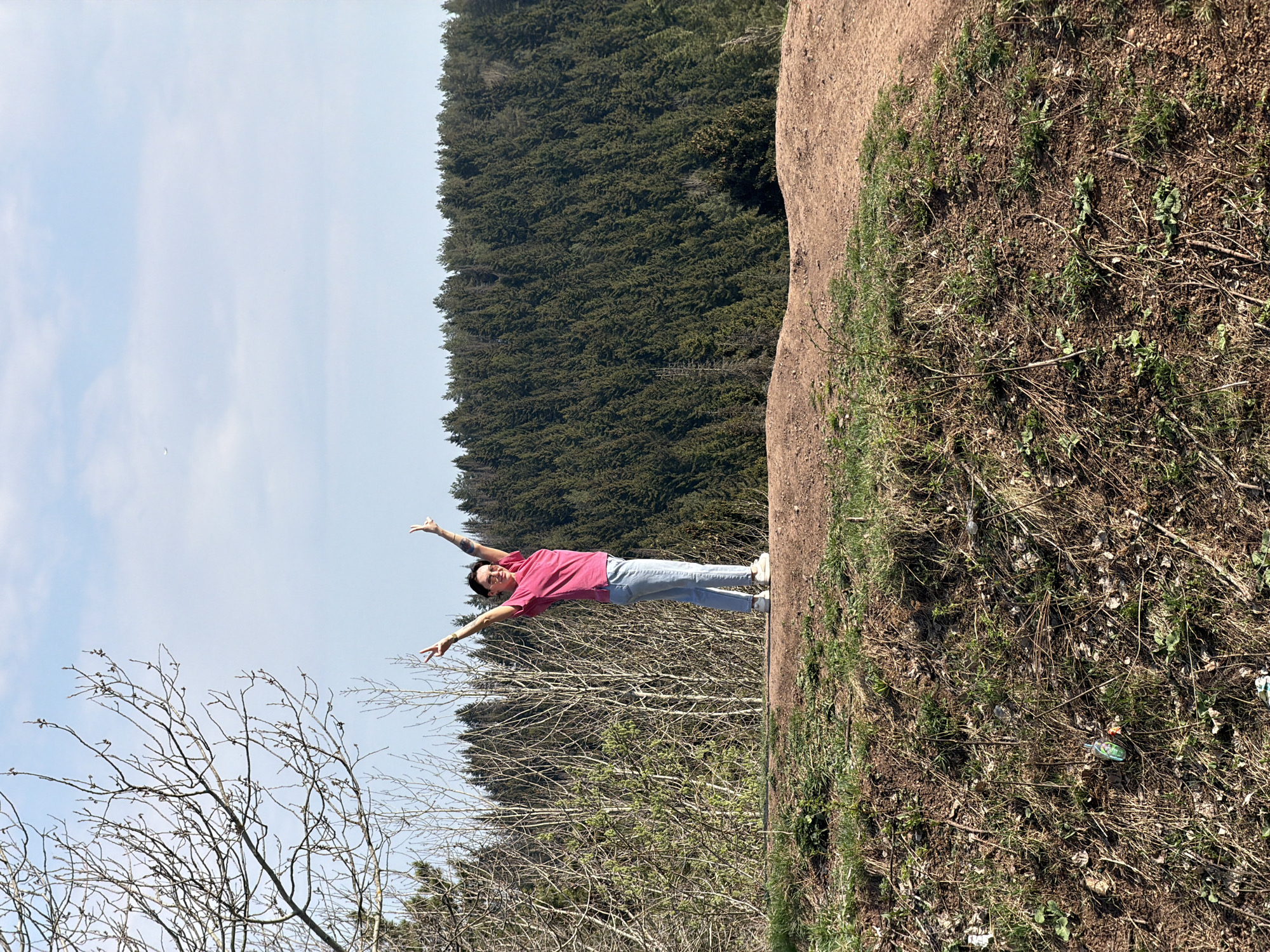 A medium shot depicts a person standing on a hillside, arms raised, with the background featuring a forest and sky. The person is wearing a pink shirt, blue jeans, and white shoes. The hillside is covered in dry vegetation and soil. The forest consists of dense evergreen trees. A leafless tree is visible in the lower left corner. The sky is light blue with some clouds. The location is likely a natural area within or near Perm, Russia, but there's no definitive confirmation of the exact location in the image itself.