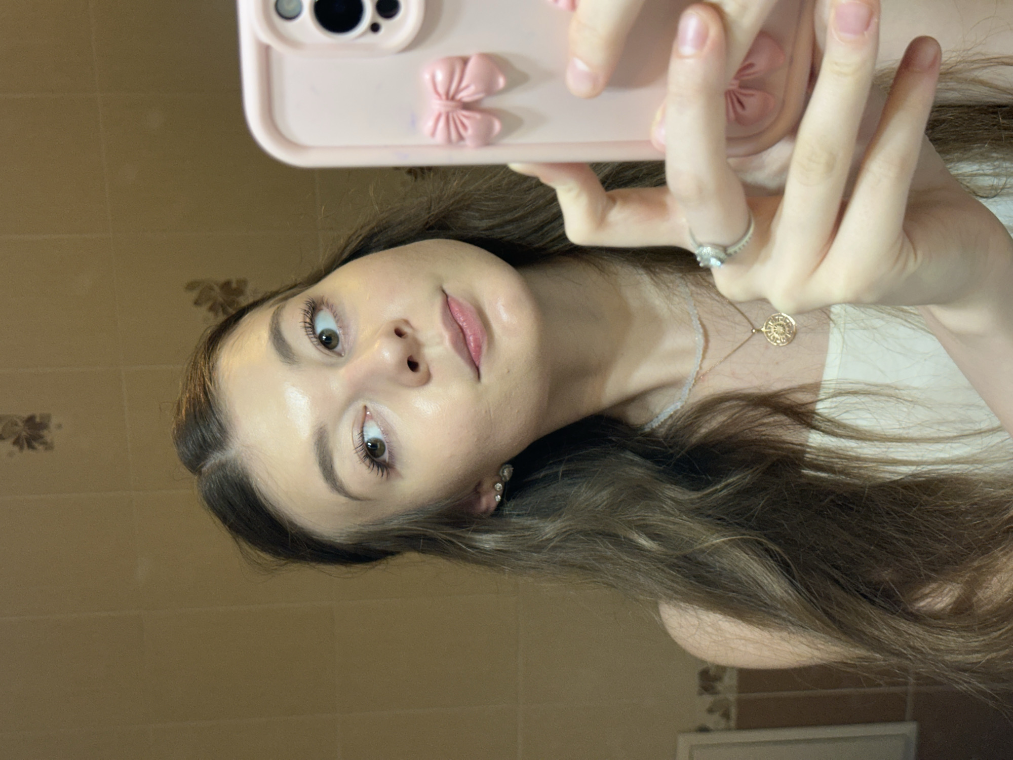 A young woman with long, wavy brown hair is taking a selfie in a bathroom. She is looking to her right, with her left eye glancing towards the camera phone. She is wearing a white top, a necklace with a circular pendant, and diamond earrings. She is also wearing a ring on her right hand, which is holding the pink phone case. The bathroom walls are tiled in a light brown color, and there is a mirror in the background reflecting the woman. The scene is well-lit, possibly by artificial light. There is no indication of the location being in the city of Kazan, Russia.
