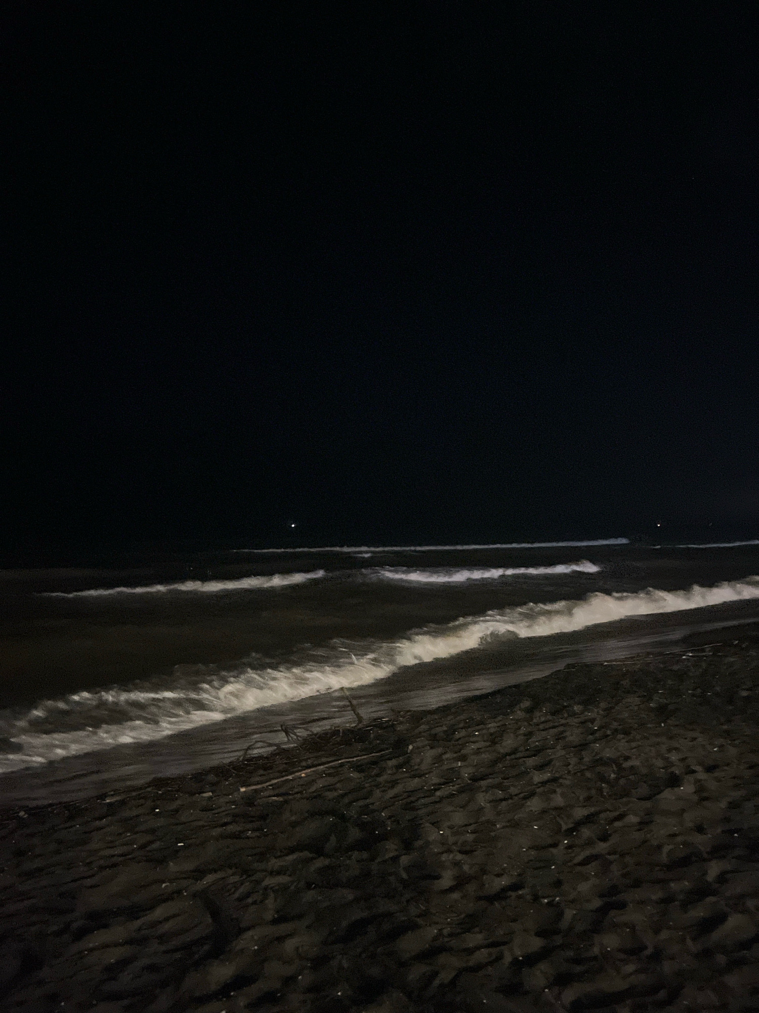 The image depicts a nighttime scene of a sandy beach with waves crashing onto the shore. The sky is dark and clear, with a single bright star or distant light visible on the horizon. The waves are illuminated by an unseen light source, creating white foam as they break. The sand on the beach is dark and appears damp, with some debris scattered across its surface. The overall impression is one of a serene and natural environment. It is not possible to determine the specific location within the city of Ureki, Georgia from this image alone.