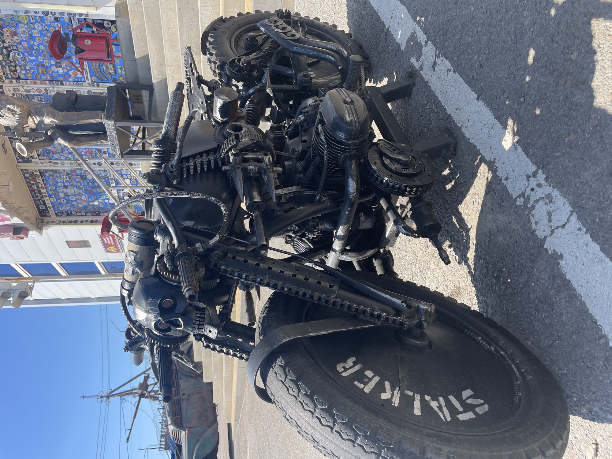 A custom-built, black motorcycle with a skull-shaped front, adorned with gears, chains, and various metal components, is parked on an asphalt surface marked with white lines. The motorcycle features a large front tire with "STALKER" painted on its side. In the background, a building with a facade decorated with numerous colorful objects and a statue of a musician playing a saxophone is visible. Stairs lead up to the building's entrance. The scene is set outdoors under a clear blue sky, suggesting a sunny day. The context of Kamensk-Shakhtinsky, Russia, indicates this likely represents a public display or an artistic installation within the city.