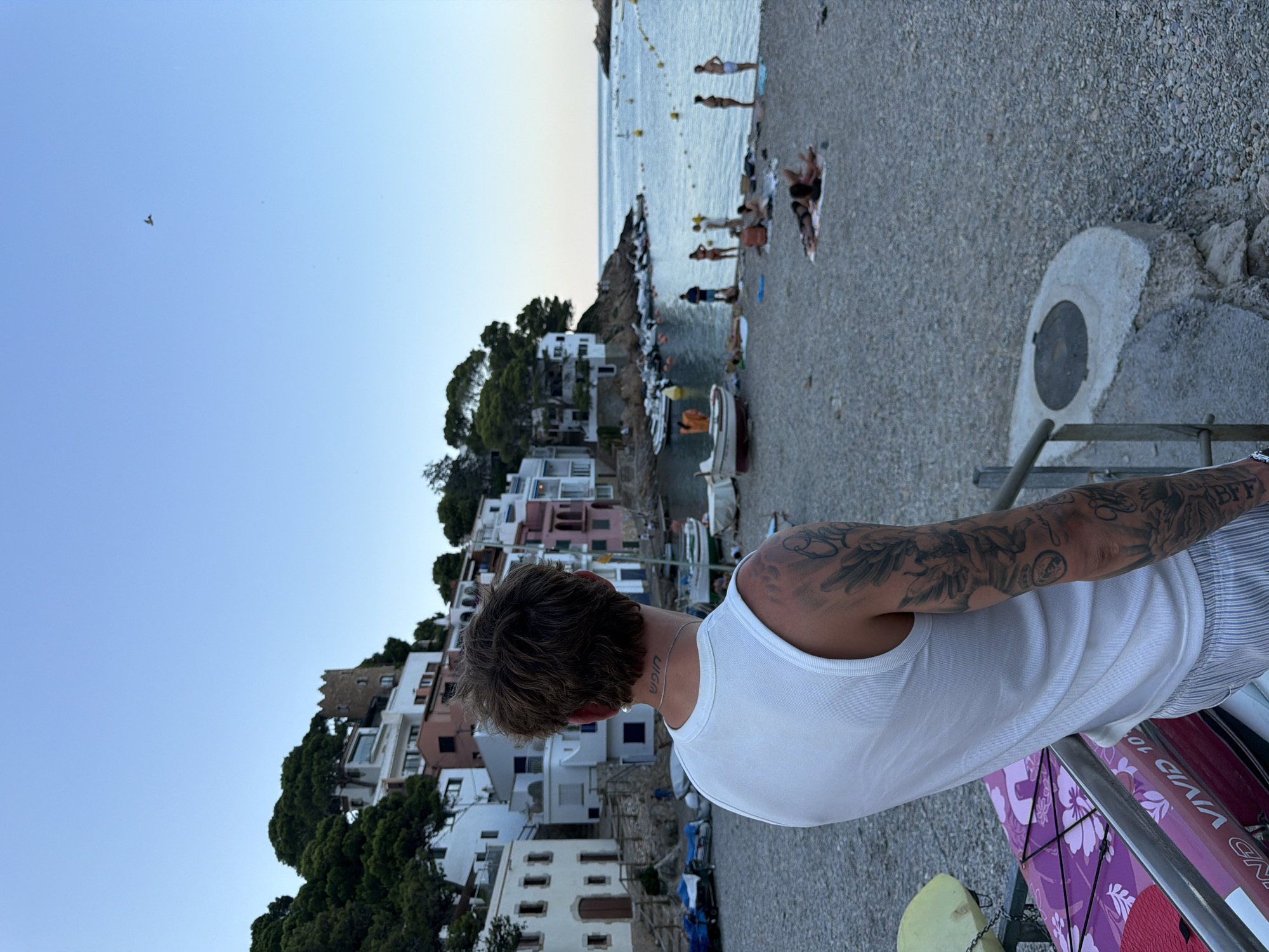 The image depicts a scenic view of the Aiguablava cove, located in the municipality of Begur, Spain. In the foreground, a young man with tattoos on his arm is seen from the back, leaning against a railing and looking out at the cove. He is wearing a white tank top and striped shorts. In front of him are parts of a purple kayak and a yellow flotation device.
The cove itself is characterized by its pebble beach, where several people are scattered, some sunbathing, others standing or walking. A collection of small boats are moored in the calm, blue water. Behind the beach, a hillside is covered with dense green trees and dotted with whitewashed houses with colorful accents, characteristic of the Mediterranean architectural style. A small, ancient-looking tower is visible on the hill, adding to the historical charm of the location. The sky is clear and blue, with a bird in flight visible in the distance. The overall atmosphere is serene and idyllic, capturing a typical summer day in this picturesque coastal area.