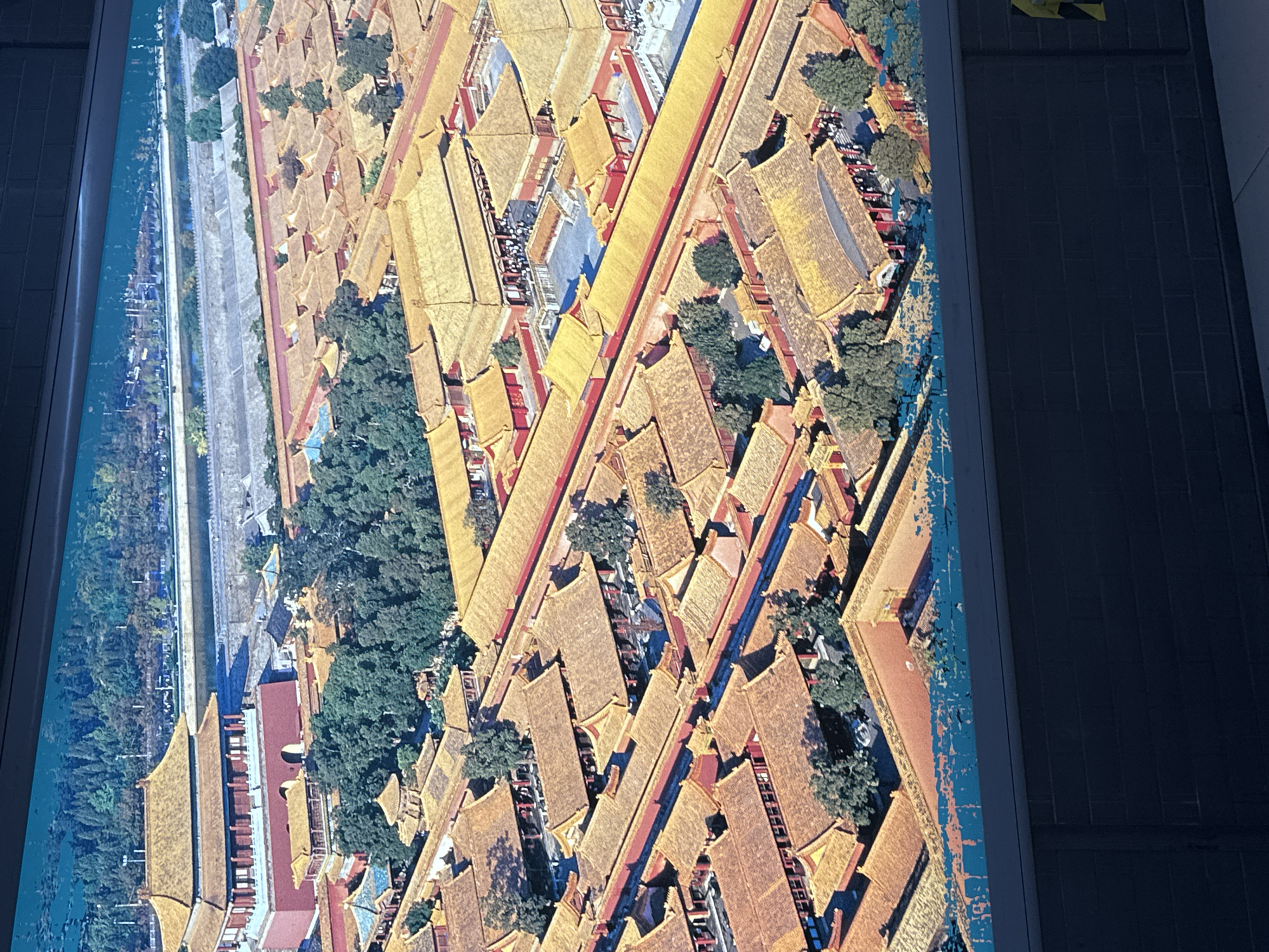 This is an aerial view of the Forbidden City in Beijing, China, specifically within the Xicheng District. The image shows a vast complex of traditional Chinese imperial palaces with distinctive golden-tiled roofs and red walls. Numerous courtyards, buildings of varying sizes, and tree-lined pathways are visible. A wide moat or river surrounds a section of the complex in the background. Several people can be seen walking in the courtyards. The overall scene is one of grand architecture and historical significance within an urban setting.