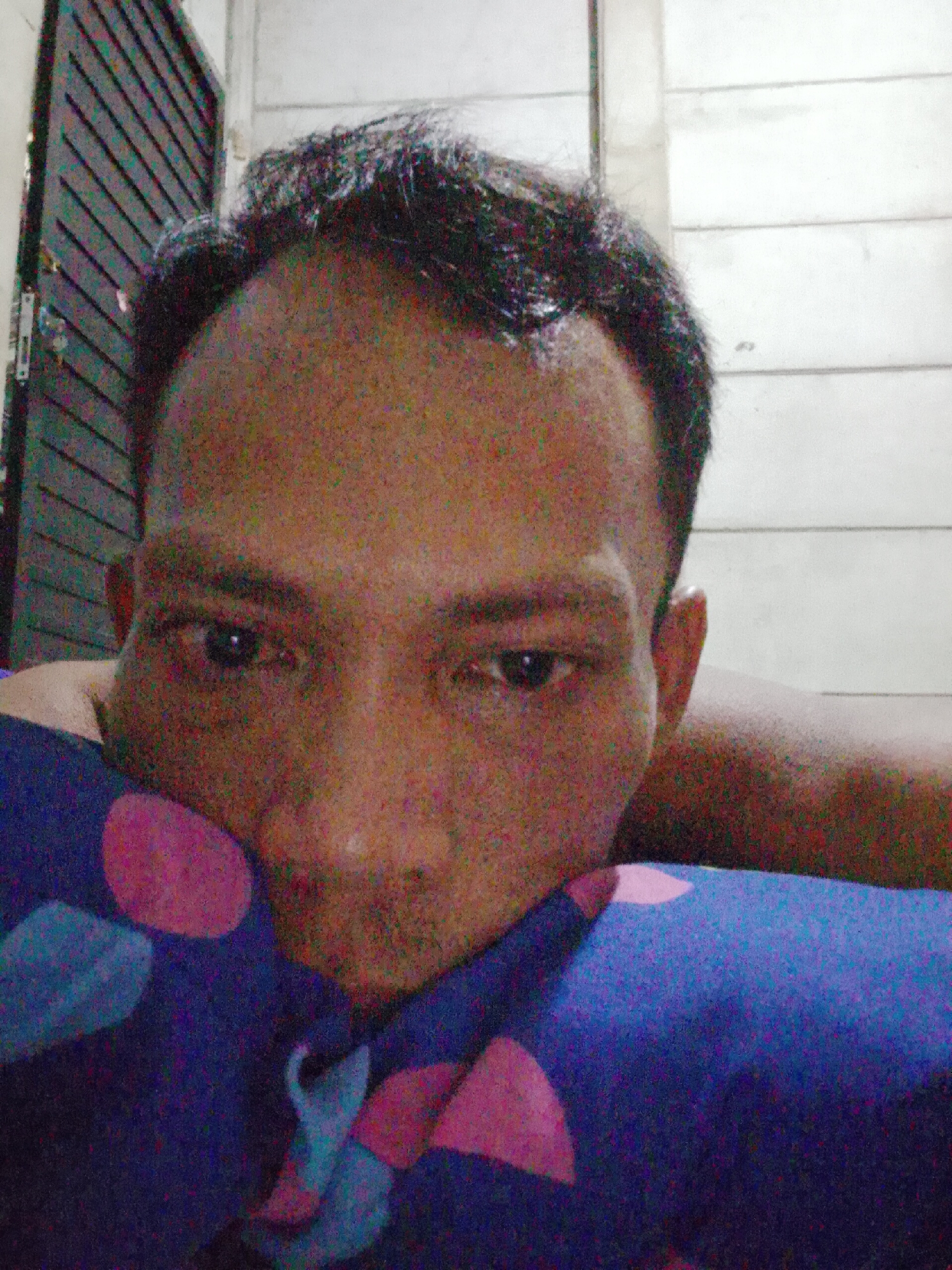 A person is lying down, resting their head on a blue pillow with pink and cyan polka dots. The person appears to be male with dark hair and skin. The background shows a white wall with horizontal lines and a dark-colored door with horizontal slats. The image was taken in Bukit Ketok, Indonesia.
