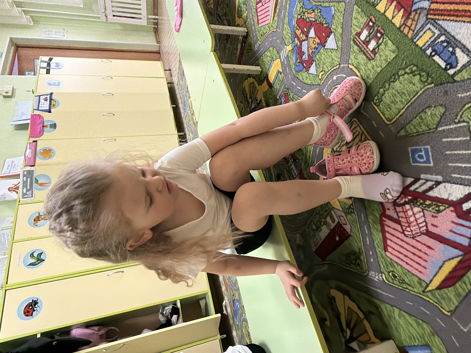A young Caucasian girl sits on a green-topped table with her legs crossed, pink sandals on her feet, and wears a white t-shirt and dark shorts. She is in a room that appears to be a children's play area or classroom, with light yellow cabinets visible along the wall behind her. Some of the cabinets have illustrations of animals on them. The girl is near a rug that has roads and building designs on it, resembling a play mat for toy cars. The photo was taken indoors. The precise location in Chaikovsky, Russia, cannot be determined from the image alone.