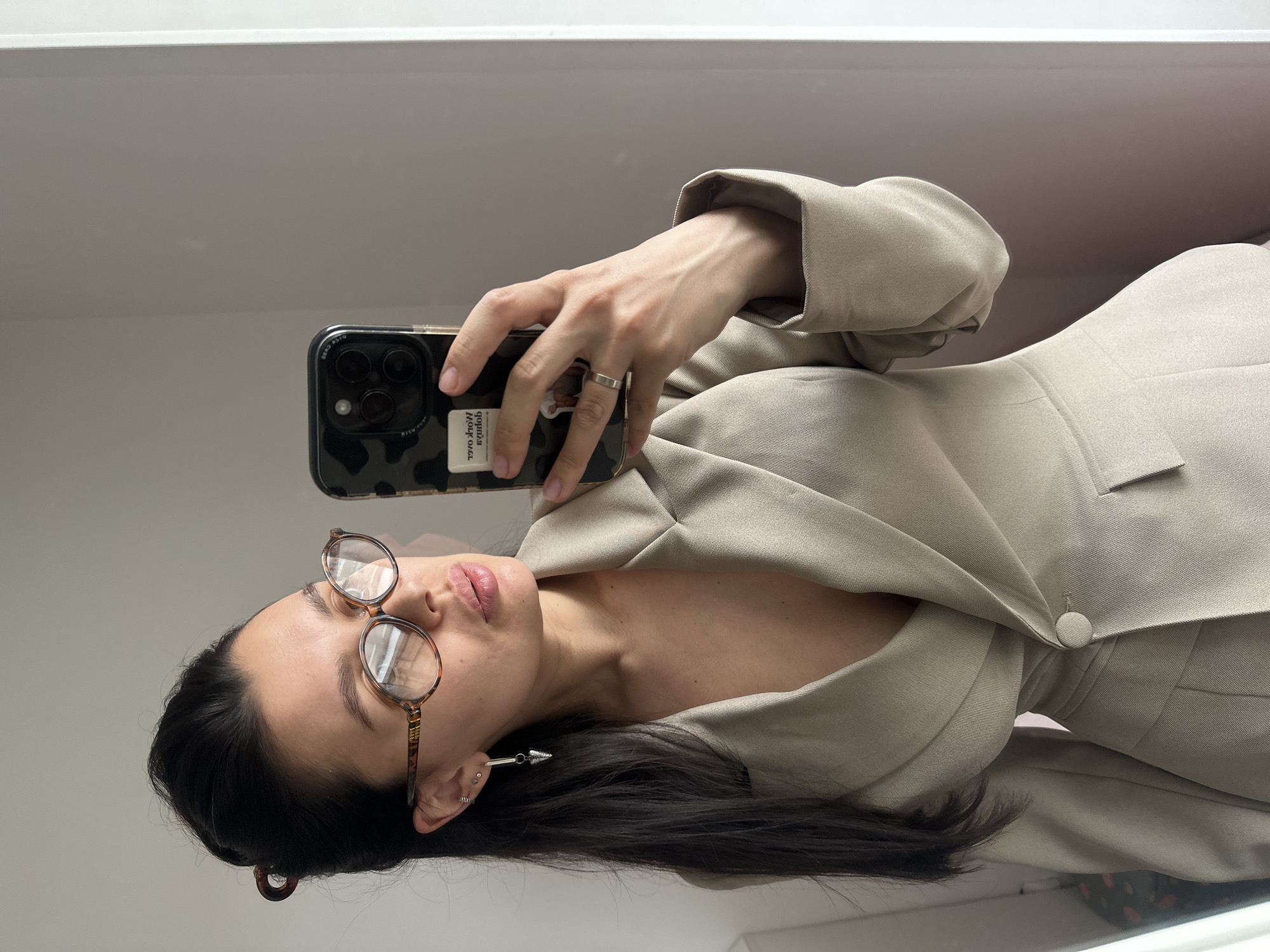 A woman with dark hair and glasses is taking a selfie. She is wearing a beige blazer. She is holding a phone with a patterned case. She has multiple piercings in her ear and is wearing a long, silver earring. The background is a plain white wall. There is no information in the image to determine a location in the city of Chaikovsky, Russia.