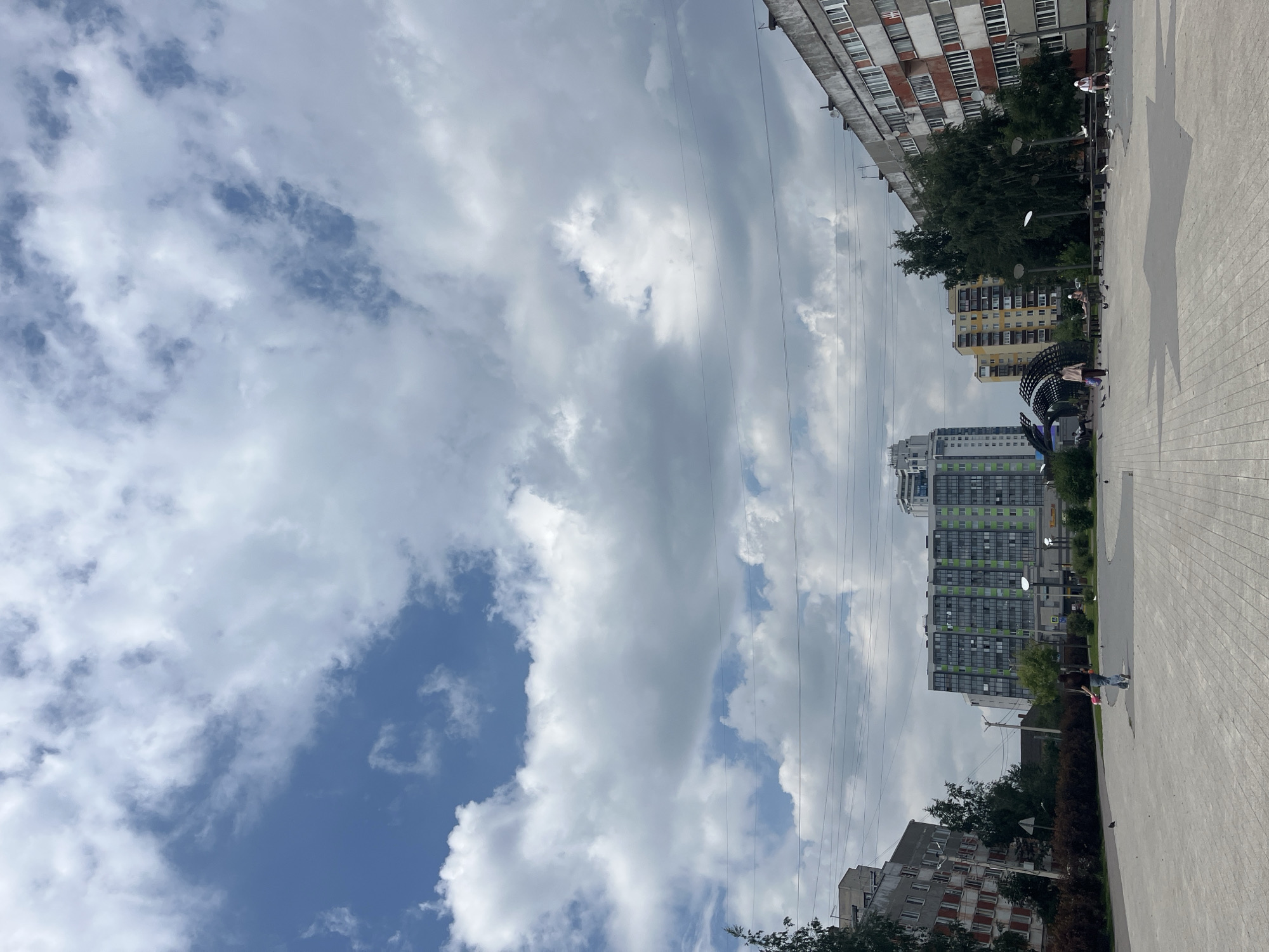 In the city of Imeni Sverdlova, Russia, a public plaza is depicted on a cloudy day. A large, modern, multi-story apartment building with a green and gray facade dominates the central background. To its left, a older, gray apartment building with red-accented windows is visible, partially obscured by trees. To the right, a weathered, red and gray apartment building with numerous windows is prominent. Several power lines crisscross the sky, stretching horizontally across the frame. The plaza itself is paved with light-colored interlocking stones, forming a large, star-shaped pattern in the foreground. A few scattered individuals are present in the plaza, including one person walking away from the camera with a pink object, and another person sitting on a bench. Birds are scattered across the plaza, some in flight and others on the ground. Trees and bushes line the edge of the plaza, separating it from the surrounding buildings.