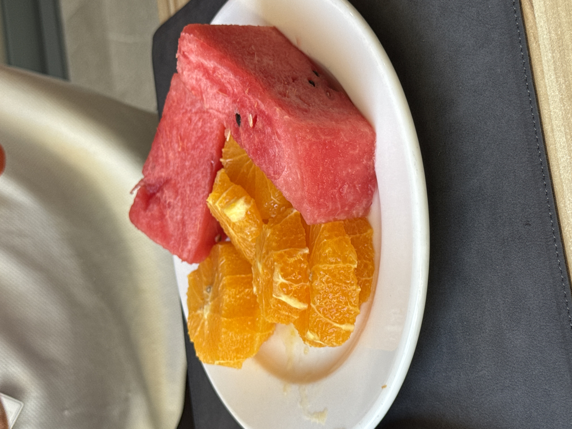 A white plate holds sliced watermelon and oranges. The watermelon is in wedge shapes, and the oranges are in circular slices, some stacked. The plate is on a dark gray placemat. In the background, a person's light-colored shirt is visible, and a wooden surface is underneath the placemat. The scene is indoors, likely in a restaurant or home. There is no information to indicate the location is Aksu, Turkey.