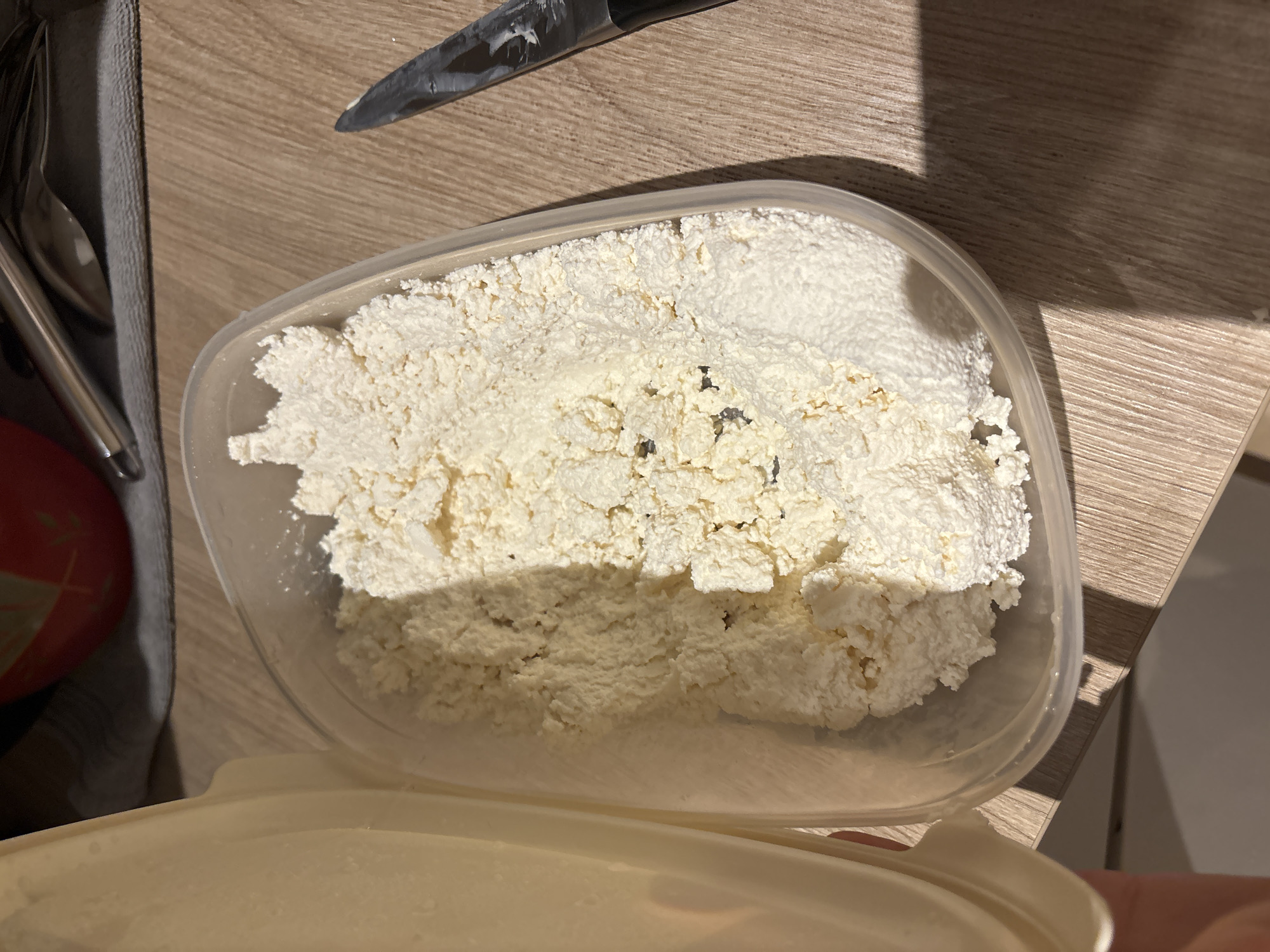 The image shows a plastic container filled with cottage cheese on a wooden surface, possibly a kitchen counter. The container is partially opened, with its lid visible at the bottom. A knife is positioned near the container's top edge. The scene appears to be indoors and there are utensils, possibly spoons and forks, near the cottage cheese. The image does not contain any information to determine location in Budva, Montenegro.