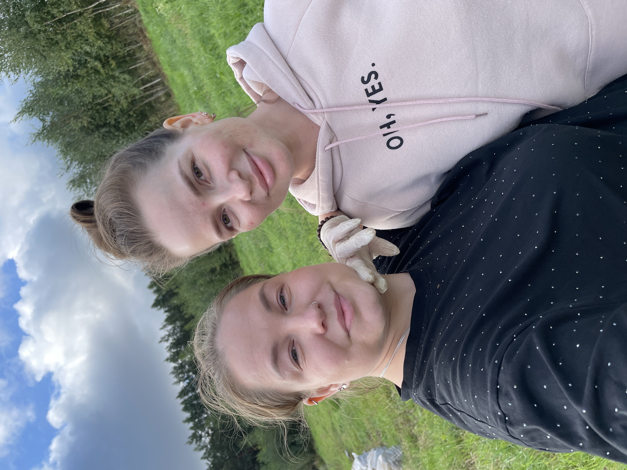 Two women are posing for a selfie outdoors, in a grassy field with trees in the background. The woman on the left is wearing a black t-shirt with small, shiny embellishments and a silver necklace. She has a nose ring and an ear piercing. The woman on the right is wearing a light pink hoodie with the text "OH, YES." printed on it. She has earrings and her hair is pulled up in a bun. One woman is wearing a light-colored glove. The sky above is cloudy. The location is identified as Baldeyka, Russia.