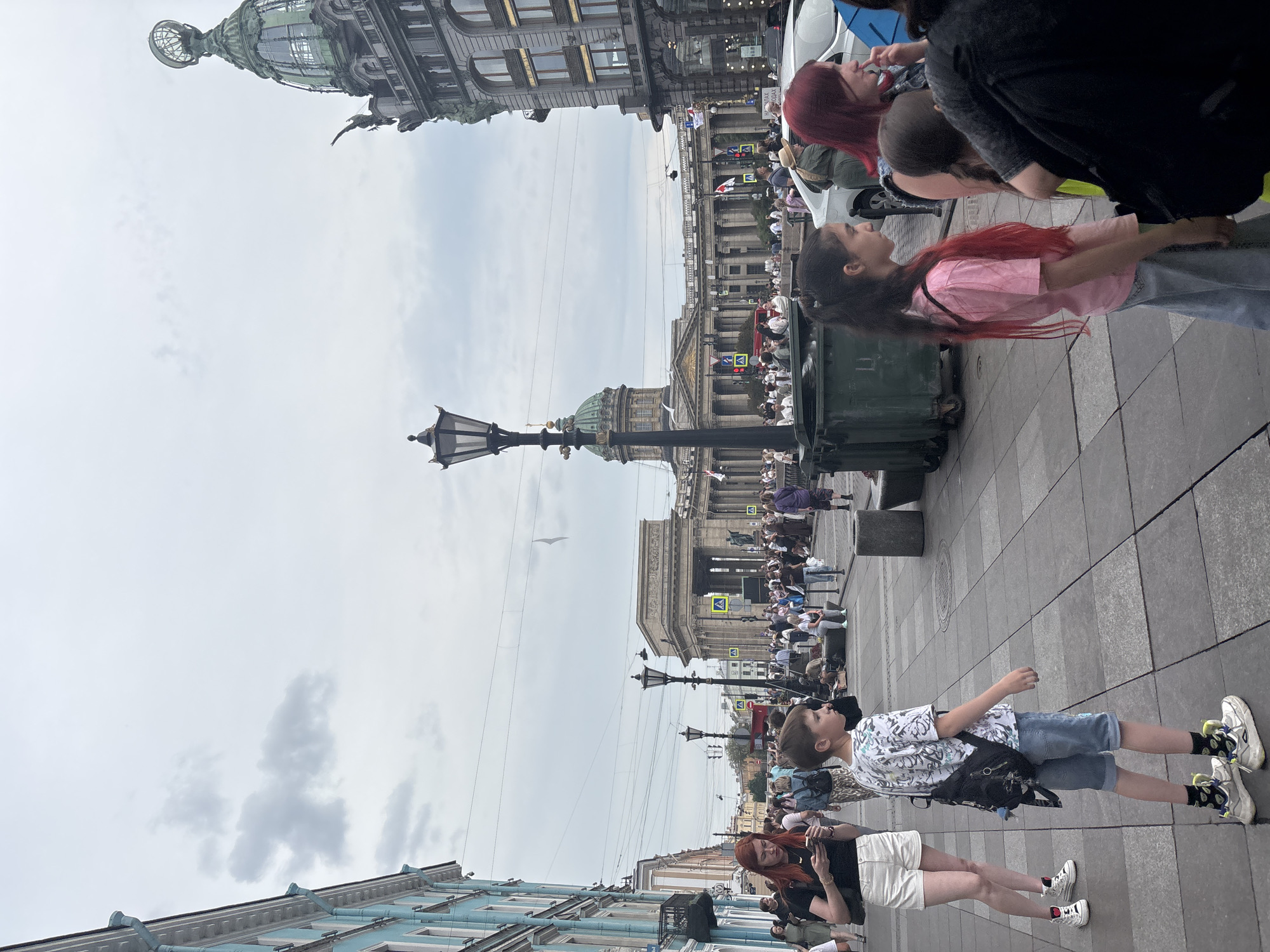 The image captures a street scene in Saint Petersburg, Russia, featuring a view towards the Kazan Cathedral, an iconic landmark with its distinctive colonnade and dome. In the foreground, a diverse group of people are walking along a paved sidewalk. A woman with red hair, wearing white shorts and a black top, is looking at her phone. Beside her, a young boy in a t-shirt and denim shorts is walking, carrying a backpack. Further to the right, a young girl with long dark hair and a pink shirt is walking, and partially visible behind her are other individuals. A large green dumpster is positioned on the sidewalk. In the background, the grand architecture of the Kazan Cathedral is prominently displayed, with a crowd of people gathered in front of it. The sky is overcast. The overall impression is of a bustling urban environment on a cloudy day.