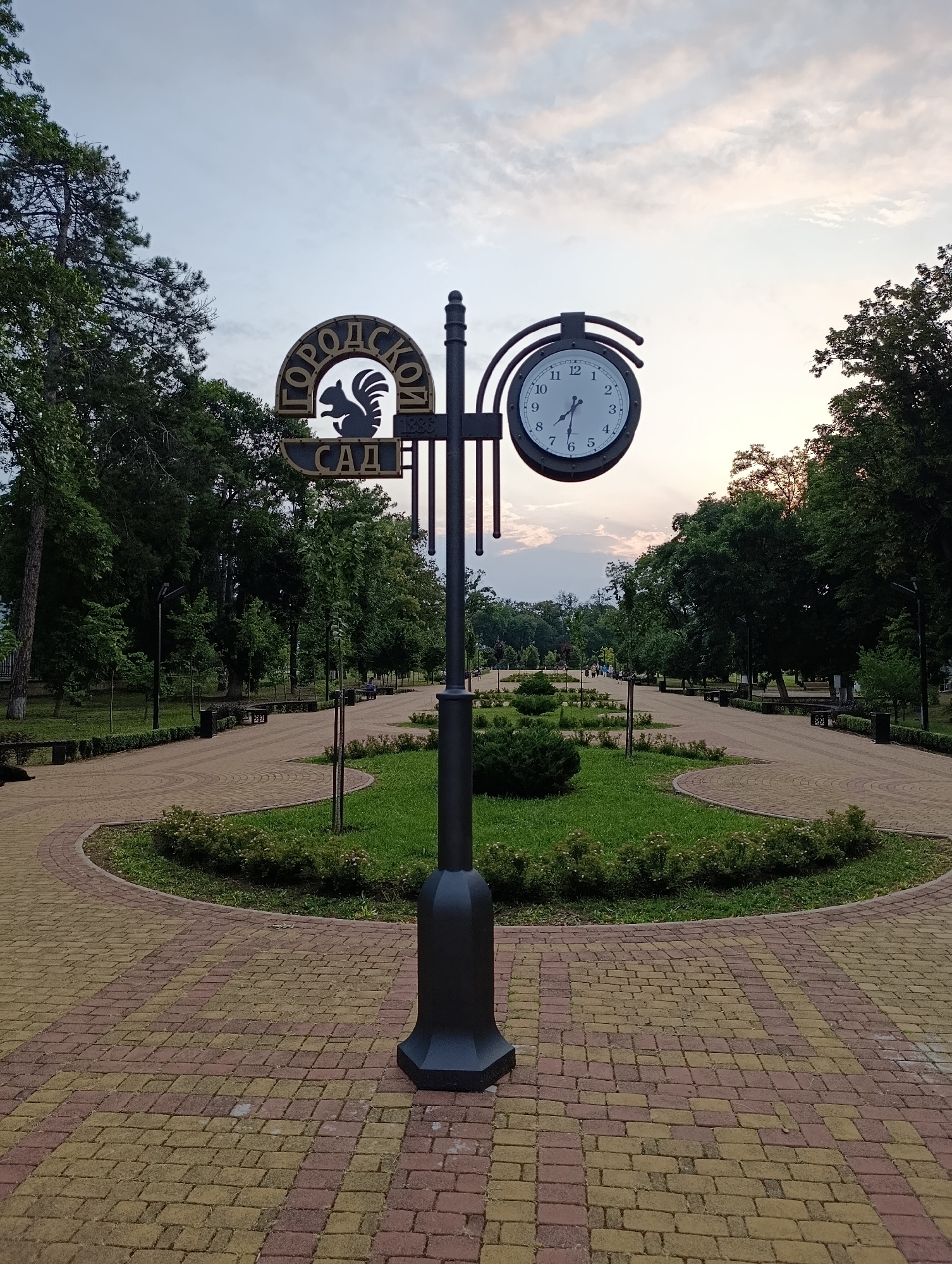 The image shows a double-sided clock mounted on a lamppost in a park. The clock face displays the time as approximately 1:45. Above the clock, there is a circular sign with the text "ГОРОДСКОЙ САД" (City Garden) and a silhouette of a squirrel. The park has paved walkways, grassy areas, benches, and trees. In the background, several people can be seen walking along the path. The sky is overcast with soft, diffused light, suggesting it may be early morning or late evening. The lamppost and sign are in the foreground, centered in the frame.