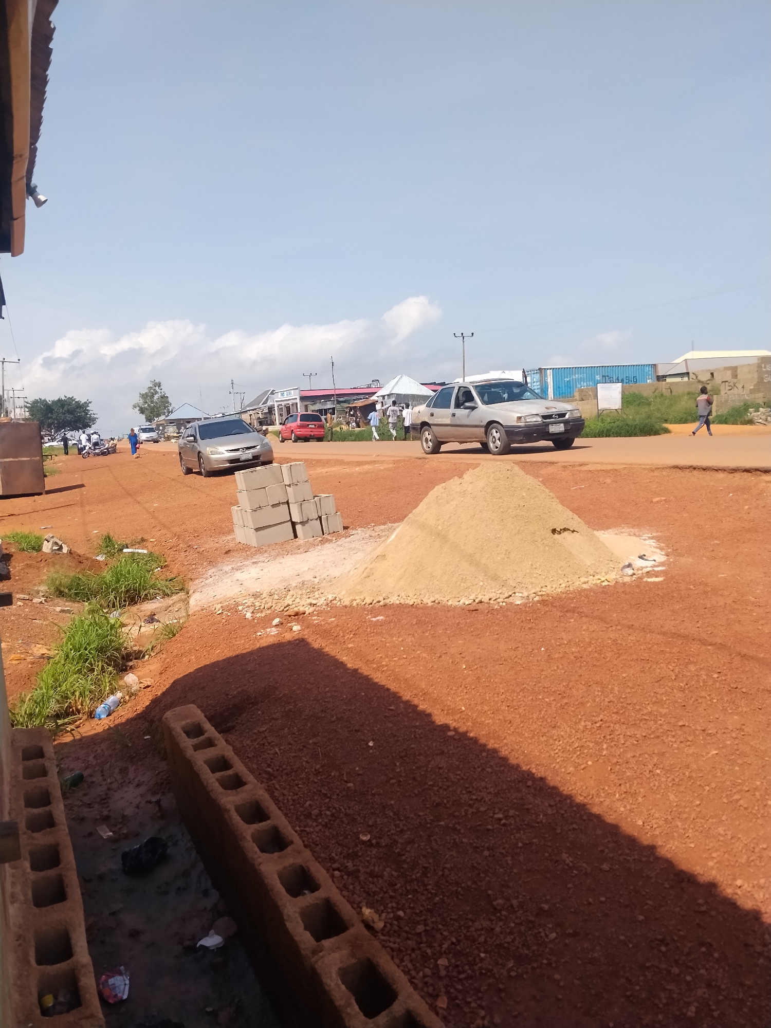 The image depicts a street scene in Durumi, Nigeria, on a clear day. The road is unpaved and appears to be under construction or repair, with a pile of sand and stacks of concrete blocks present. Several vehicles, including a silver sedan and a red hatchback, are on the road. In the background, buildings line the street, and some individuals are visible walking or standing. A blue shipping container is also noticeable in the distance. The scene suggests a typical urban environment in Nigeria, possibly a commercial or residential area undergoing infrastructure development.