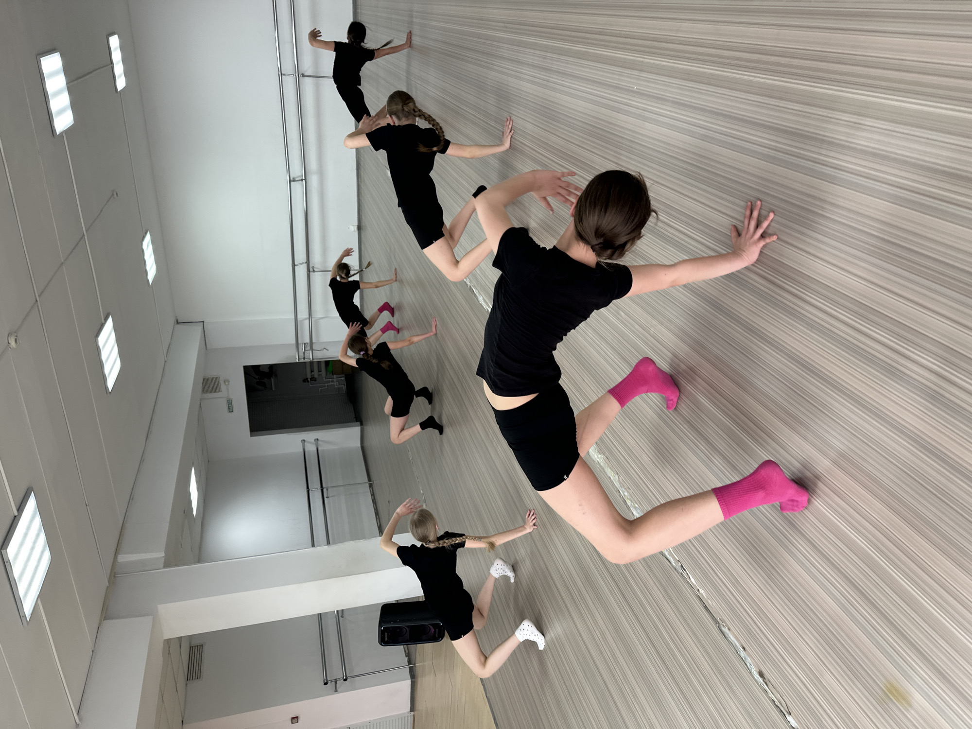 The image shows a group of young dancers in a dance studio. They are dressed in black tops and shorts, with some wearing socks or shoes. They are positioned against a wall with a striped pattern, seemingly practicing a choreographed movement with their arms outstretched. The room has white walls and bright lighting fixtures on the ceiling. The location appears to be a dance studio in Votkinsk, Russia.