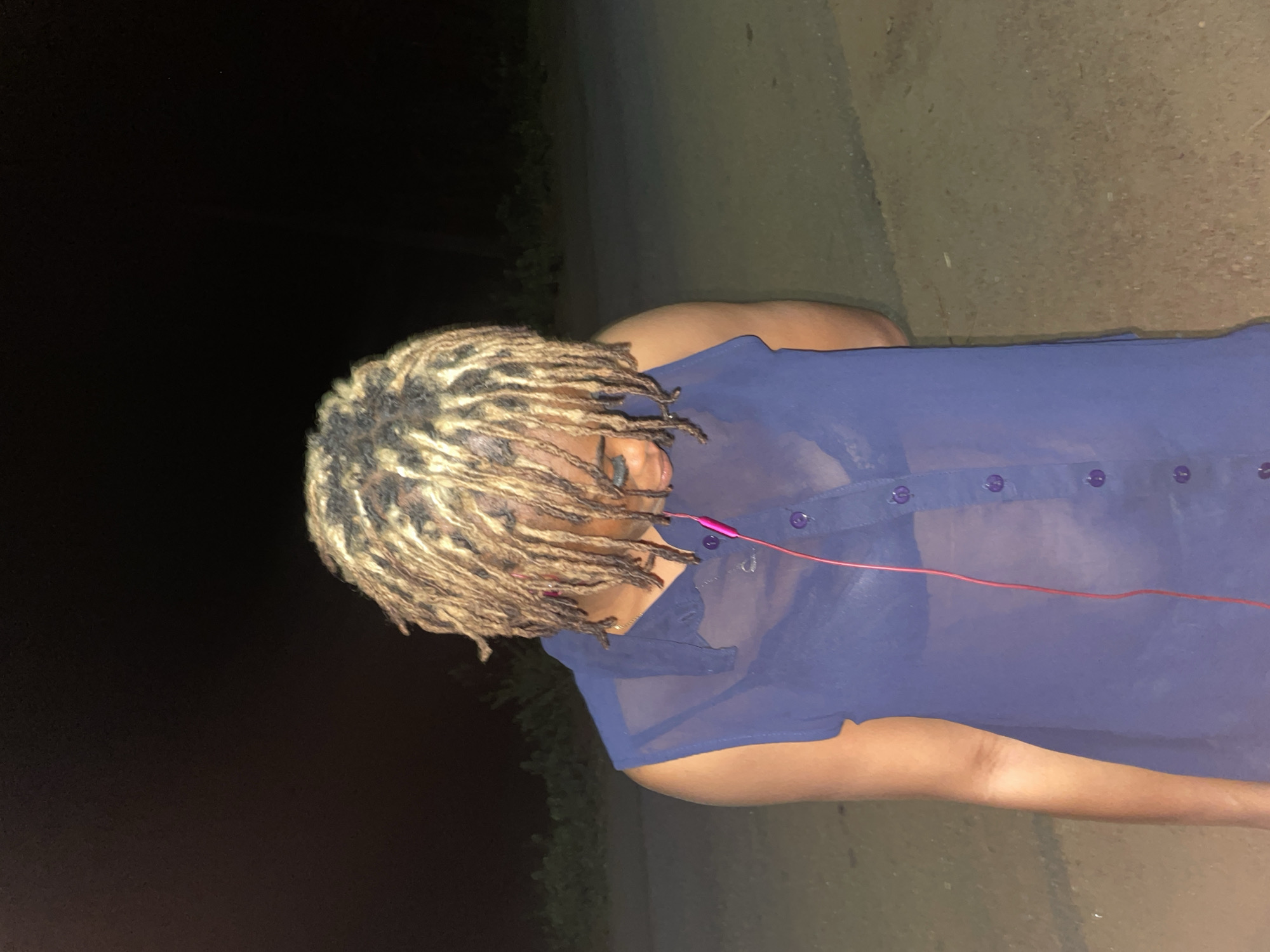 The image depicts a person with blonde dreadlocks wearing a sheer blue button-down shirt and red headphones. The person is outdoors. The background is divided between darkness on the left and a blurry grey area suggesting a path or road on the right. There is no definitive evidence within the image to confirm its location in Ogbe, Nigeria. The scene is poorly lit.